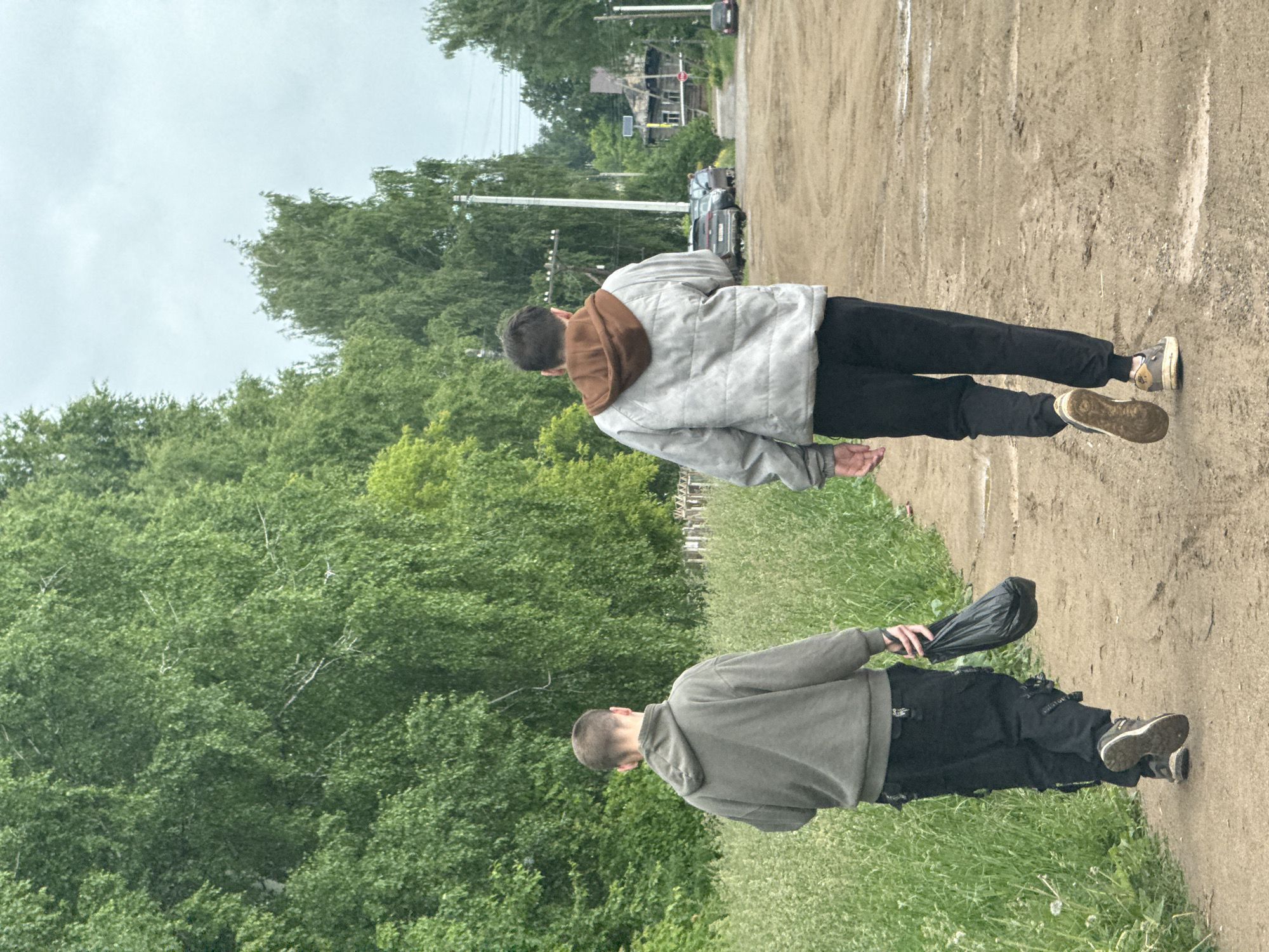 The image depicts two young males walking away from the viewer on a wide, unpaved dirt path. The setting is an outdoor, rural or semi-rural area in Фоки, Russia, under an overcast, light grey sky.
The person in the foreground wears a grey hoodie, dark pants with visible buckles or straps, and dark sneakers with white soles. They are carrying a black bag in their right hand. Further down the path, the second person wears a light grey jacket with a brown hood, black pants, and lighter-colored sneakers. Both individuals have short, dark hair.
To the left of the path, dense green foliage and trees dominate the landscape. To the right, the path extends into a village street where utility poles and power lines are visible. Several cars are parked along this street, and in the distance, older wooden houses with dark roofs can be seen amongst the trees. A blurry red circular sign, possibly a stop sign, is faintly visible near one of the distant houses. The dirt path itself shows tire tracks and is bordered by patches of green grass. The activity captured is simply two individuals walking through this rustic environment.