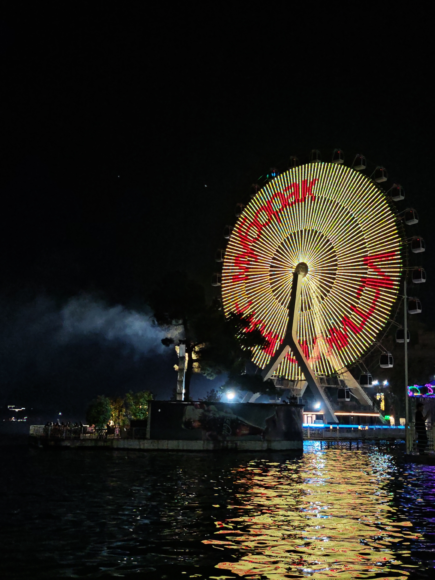 This is a nighttime photograph of a brightly lit Ferris wheel in Dushanbe, Tajikistan. The Ferris wheel dominates the right side of the frame, its spokes and carriages outlined in yellow and red lights. The lights on the Ferris wheel form a pattern, with red lights spelling out Chinese characters. Below the Ferris wheel, a body of water reflects the colorful lights, creating a shimmering effect. In the middle ground, a dark, elongated structure, possibly a boat or pier, is visible, with people gathered on its surface. To the left of this structure, a plume of white smoke rises into the dark sky, partially obscuring some of the lights on the Ferris wheel. The overall scene suggests an amusement park or waterfront entertainment area at night.