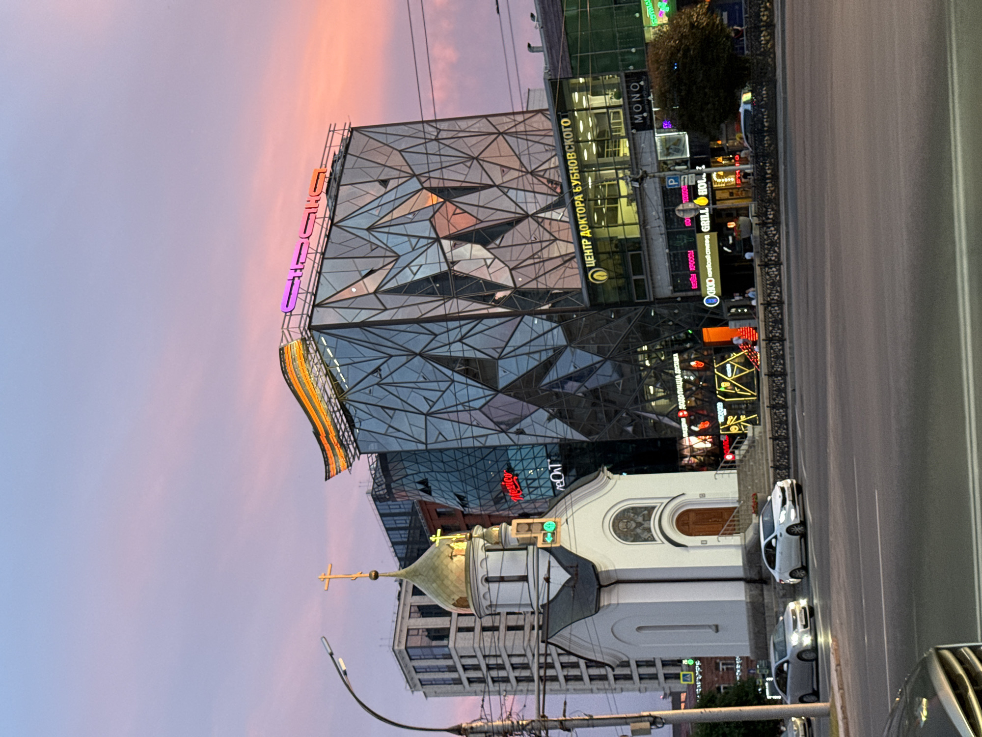 This image, taken in Novosibirsk, Russia, depicts a city street scene at dusk, characterized by a pastel pink and purple sky. The foreground features a modern building with a distinctive geometric glass facade, adorned with colorful neon signs. To the left of this building, a small, white church with a golden dome and a cross stands in contrast to the contemporary architecture.
The street itself is visible on the right, with cars parked and driving on it. Various signs are displayed on the buildings, including "MONO," "ЦЕНТР ДОКТОРА БУБНОВСКОГО" (Center of Doctor Bubnovsky), and "ДОДО ПИЦЦА" (Dodo Pizza), indicating commercial establishments. The overall atmosphere suggests a lively urban environment transitioning into evening. The lighting from the neon signs and streetlights illuminates the scene, creating a blend of natural and artificial light. The image seems to capture a moment of urban life as day turns into night.