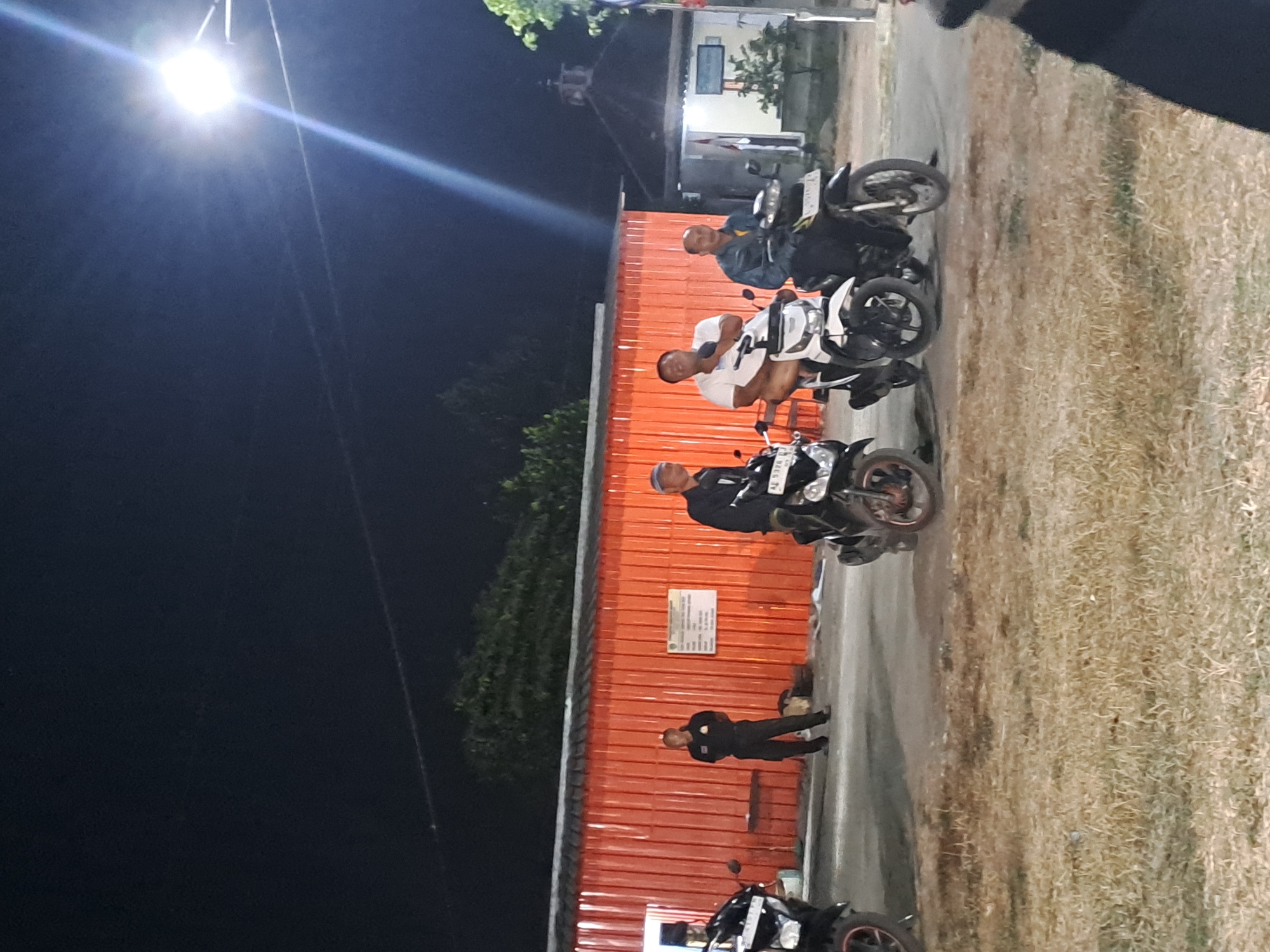 The image depicts a nighttime scene in front of an orange corrugated metal building. Three men are seated on motorcycles. The man on the left is wearing a white t-shirt, and the man on the right is wearing a dark jacket and a white cap. To the left of the men, a person in uniform, possibly a security guard, stands next to a motorcycle. The building has a white sign with text on it. In the background, there is a smaller building with a steeple and a flag. The ground in the foreground is covered with dry grass. A bright street light illuminates the scene from the upper right. The overall impression is a gathering of people on motorcycles at night, possibly in a commercial or industrial area of Sumberejo, Indonesia.