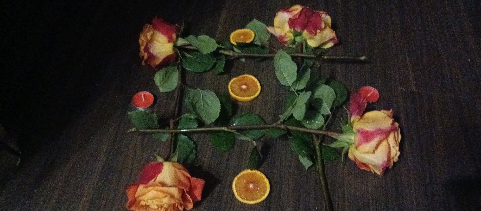 On a dark wooden surface, a square is formed using four rose stems. Each stem has at least one rose and several green leaves. Within the square, there are three orange slices, and outside the square, two small red candles are positioned on opposite sides. The setting appears to be indoors due to the wooden surface and lack of natural light. The location in the city is unknown.