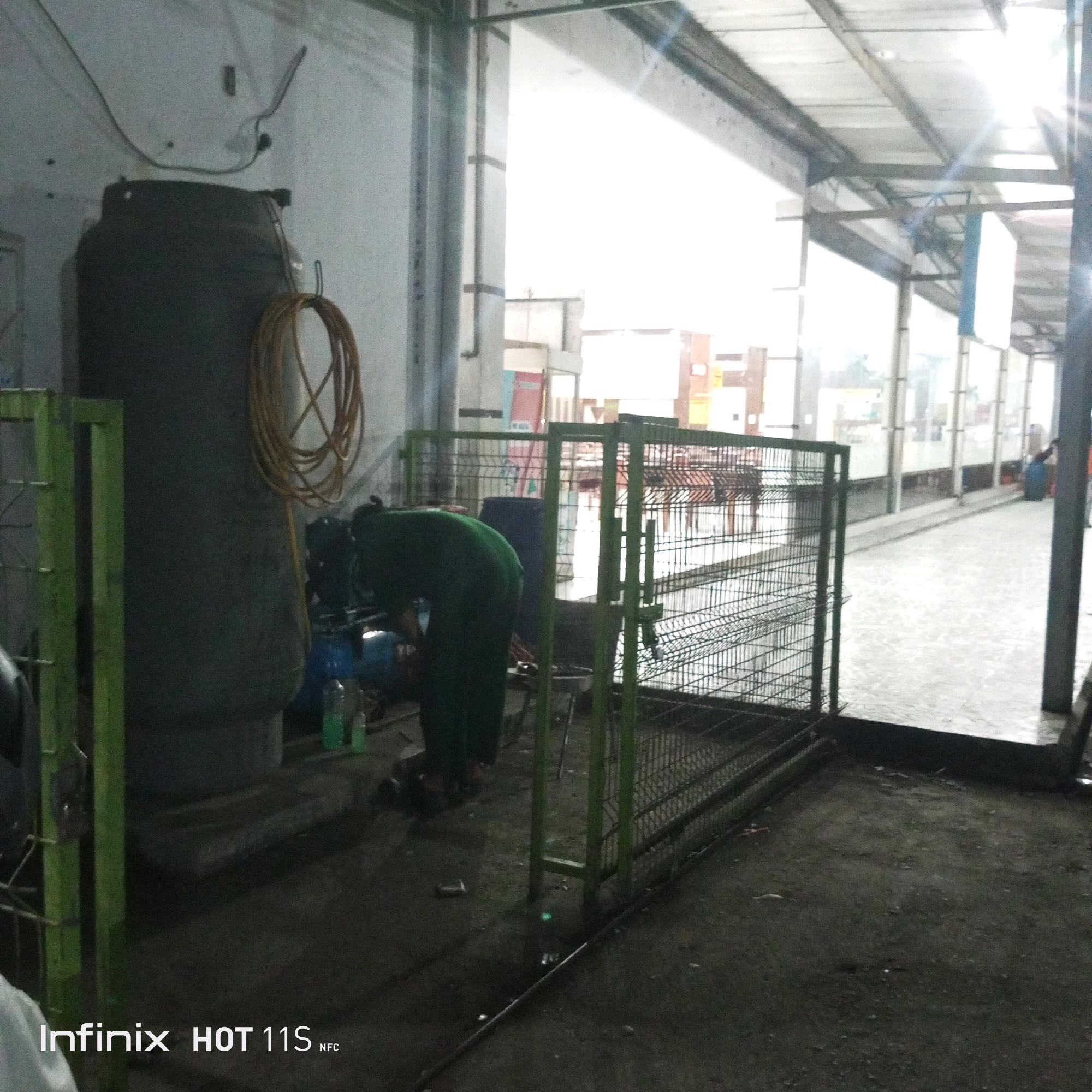 A person wearing a green uniform is working near a large, dark-colored tank. The tank has a coiled orange hose attached to it. To the left of the tank is a green metal fence. In front of the person is a blue tank and some bottles. The ground is covered in dirt and debris. The scene appears to be an outdoor or semi-outdoor industrial area with a covered walkway in the background. The image is also watermarked with "Infinix HOT 11S NFC".