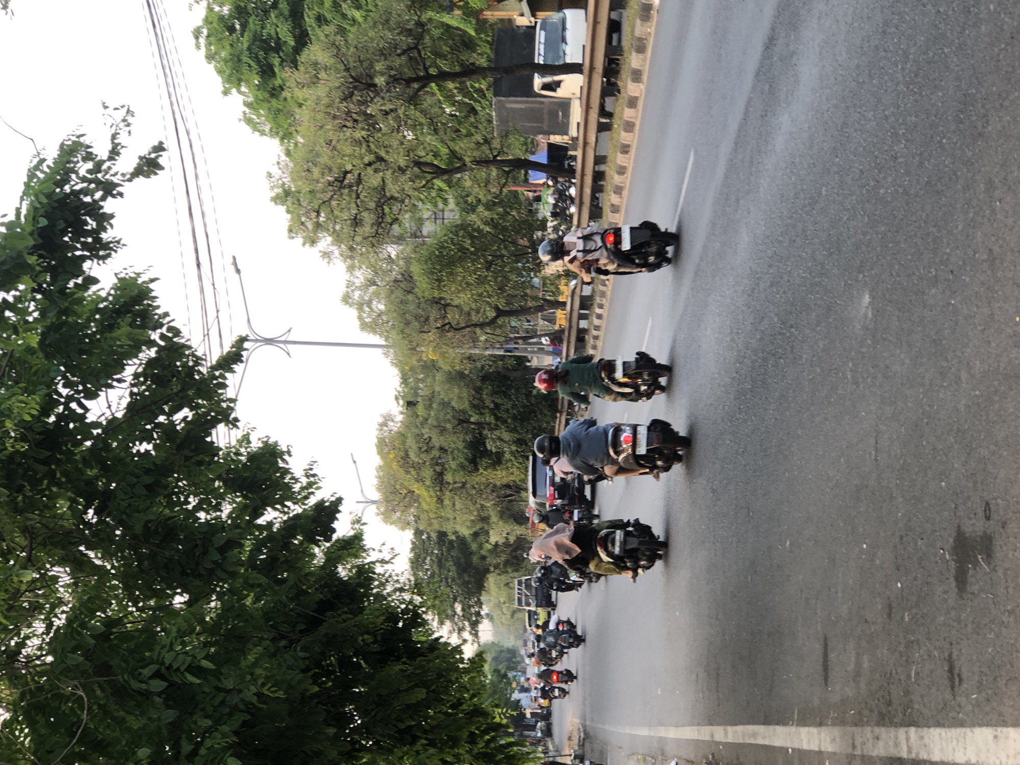 The image depicts a street scene in Kludan, Indonesia, with a focus on a busy road filled with motorcycles and some cars. In the foreground, several motorcycles are traveling along the asphalt road, with riders wearing helmets. One motorcycle in the center foreground has a rider wearing a headscarf and a dark jacket. To the right, another motorcycle rider is visible from behind, wearing a light-colored shirt and a helmet.
The road is bordered by a curb and a low wall, beyond which is a line of trees. The trees are lush and green, with some foliage extending over the road from the left side of the image. In the background, more trees are visible, along with power lines and a street light. There are also several vehicles in the distance, including a truck, a car, and a white van. The sky is overcast, suggesting it might be early morning or late afternoon. The overall impression is that of a typical urban or suburban street with active traffic.