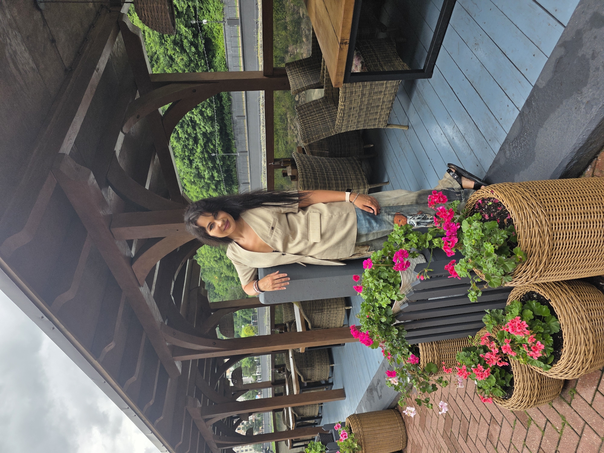 A woman stands on a wooden deck under a covered patio. She is leaning against a dark gray pillar, with her left arm resting on it. The woman has long, dark hair and is wearing a light-colored vest over a v-neck top and patterned pants. In front of her are several wicker baskets filled with pink and green plants. The deck has blue painted wooden planks. Behind the woman, tables and chairs made of wicker are visible. Beyond the covered patio, a body of water and lush green trees can be seen. The sky is overcast. The scene appears to be an outdoor seating area of a restaurant or cafe located in Estosadok, Russia, possibly along a waterfront.