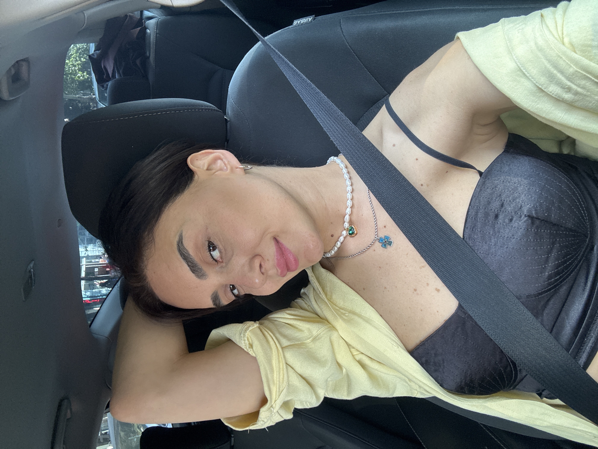 A woman with dark hair and fair skin is sitting in the driver's seat of a car, resting her right arm behind her head. She is wearing a yellow open shirt over a black bustier, a pearl necklace with a green pendant, and a silver necklace with a blue cross pendant. A black seatbelt is across her chest. The car interior is visible, including a black headrest with an "AIRBAG" label. Outside the car, through the windows, a city street scene with buildings and trees can be seen. The scene appears to be in Tbilisi, Georgia, based on the visible streetscape and architecture.