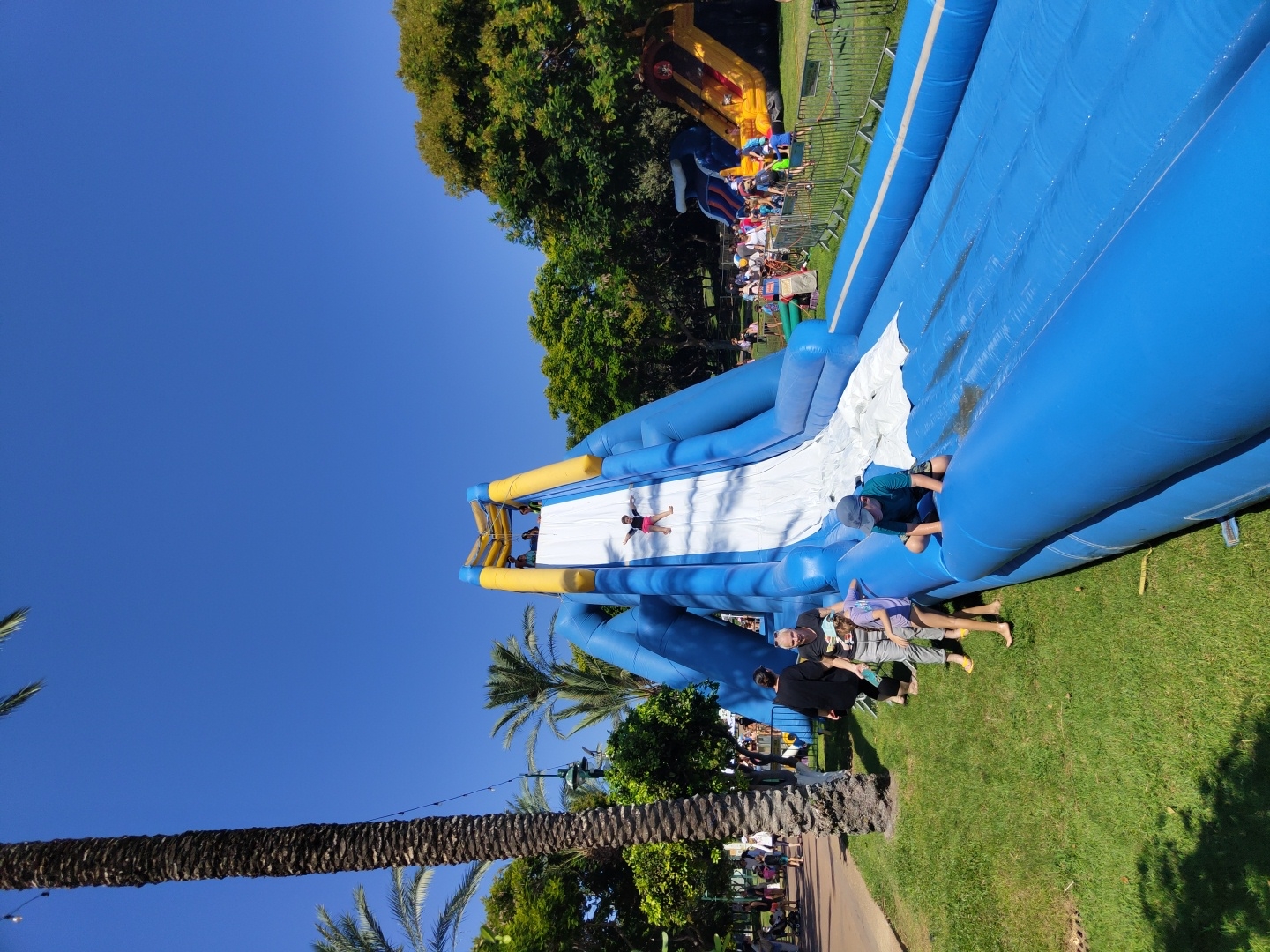 In Raanana, Israel, an inflatable water slide is set up on a grassy area on a sunny day. The slide is primarily blue with yellow accents and features a long, slippery descent. Several people are engaged with the slide. A child is sliding down the white surface of the slide, and another child, wearing a blue hat and a dark shirt, is on the lower part of the slide, appearing to be moving forward. In the background, other inflatable structures, including a yellow and orange slide, are visible, along with a crowd of people, some of whom are walking and observing. Palm trees and other foliage surround the area, and a clear blue sky is overhead.