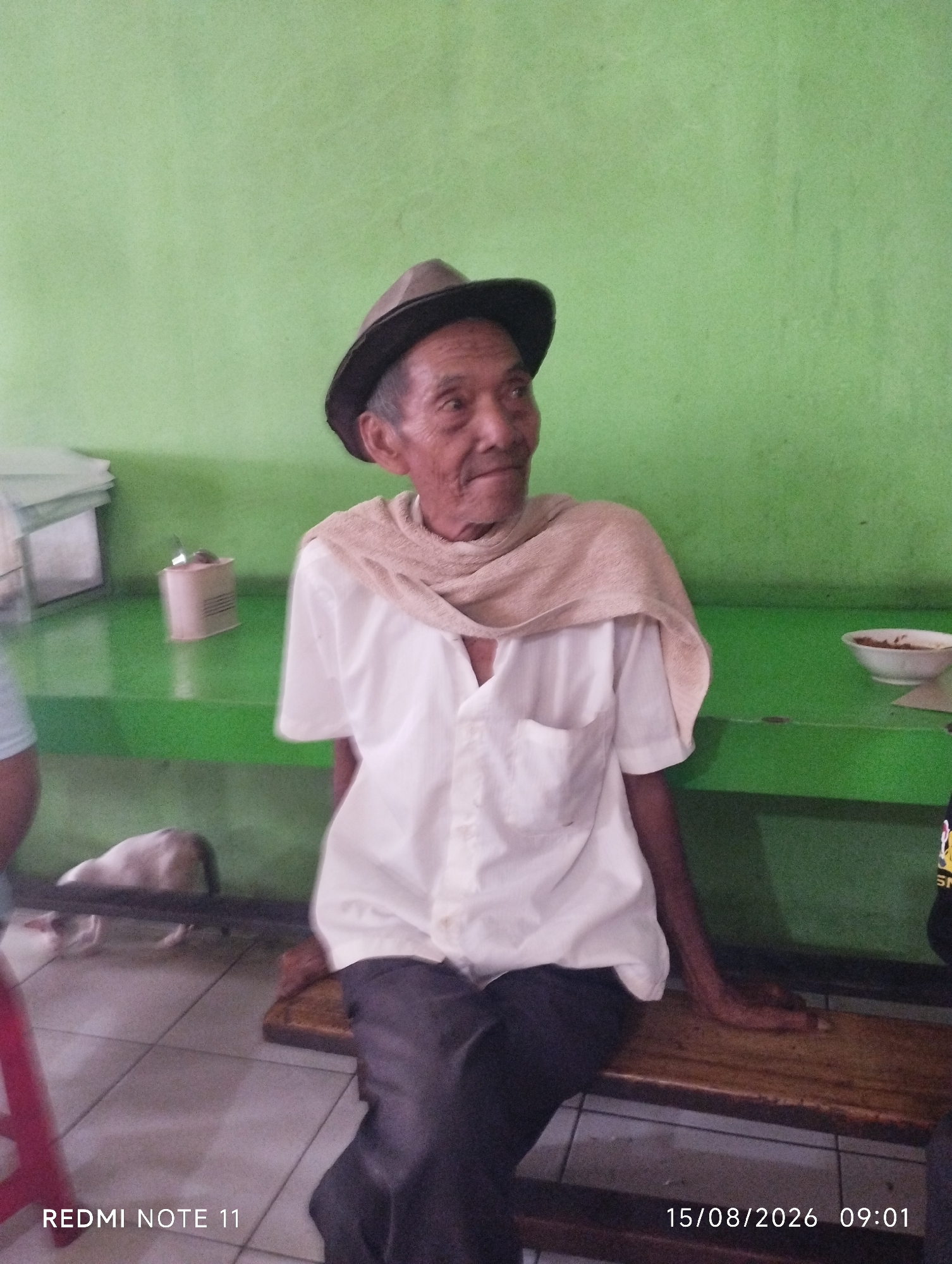 The image shows an elderly man wearing a hat, a white collared shirt, and dark pants, sitting on a wooden bench. A light-colored scarf is draped around his neck and shoulders. He is looking to his right. The background features a bright green wall. A green shelf is visible on the wall, holding various objects, including what appears to be a container and a bowl of food. In the foreground, a tiled floor is visible, and part of a red chair is on the left side of the frame. There is a dog in the background, partially obscured. The lower right corner of the image contains a timestamp and text that reads "REDMI NOTE 11 15/08/2026 09:01". The scene appears to be indoors, possibly a simple cafe or eatery. Given the information present in the image, it is not possible to determine the specific location within the city of Karawang, Indonesia.