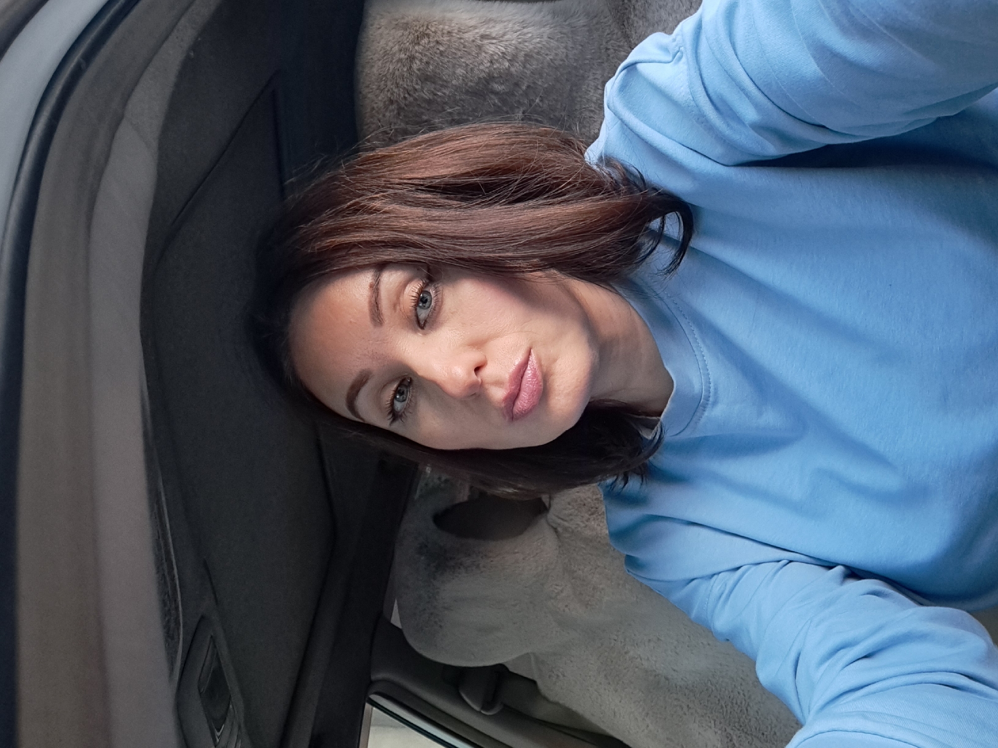 A woman is taking a selfie inside a car, with a furry seat cover visible behind her. She has brown hair and is wearing a light blue sweatshirt. The car appears to be parked, with the interior ceiling and seats in focus. The location of this image in the city of Irkutsk, Russia, cannot be determined from the visual information provided.