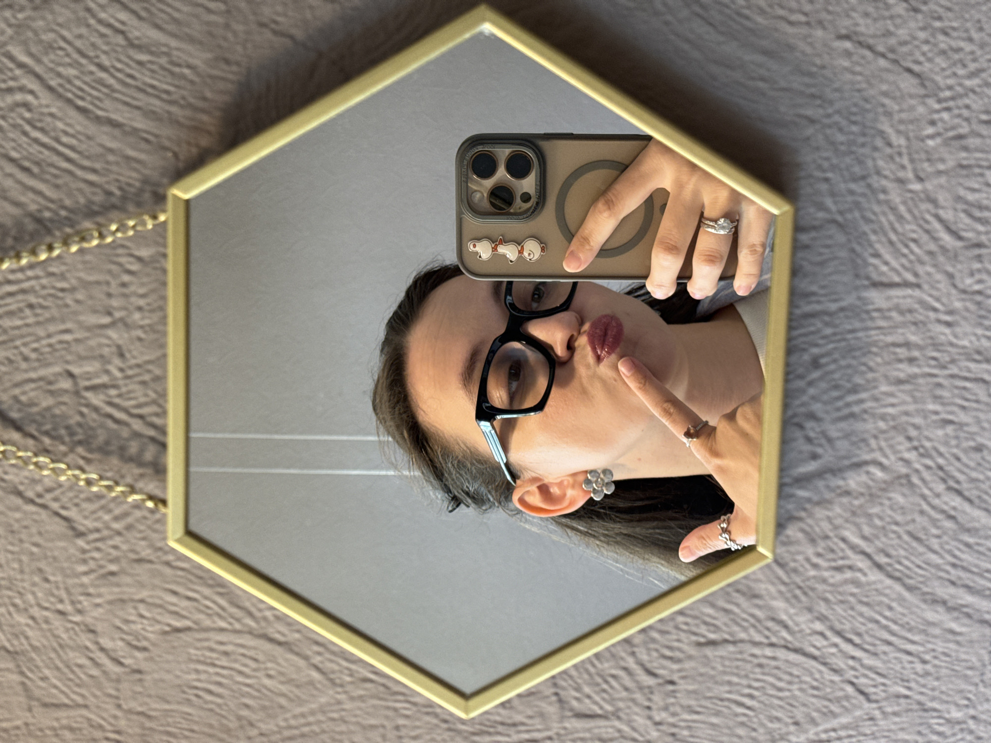 A young woman with long dark hair, wearing black-rimmed glasses and a white top, takes a selfie in a hexagonal mirror with a gold frame. She is wearing a flower-shaped earring on her left ear and two rings on her right hand. Her phone, decorated with three small duck stickers, is held up to take the photo. She is puckering her lips and has her index finger raised to her lips. The background is a textured grey wall. The location is identified as Chaikovsky, Russia.