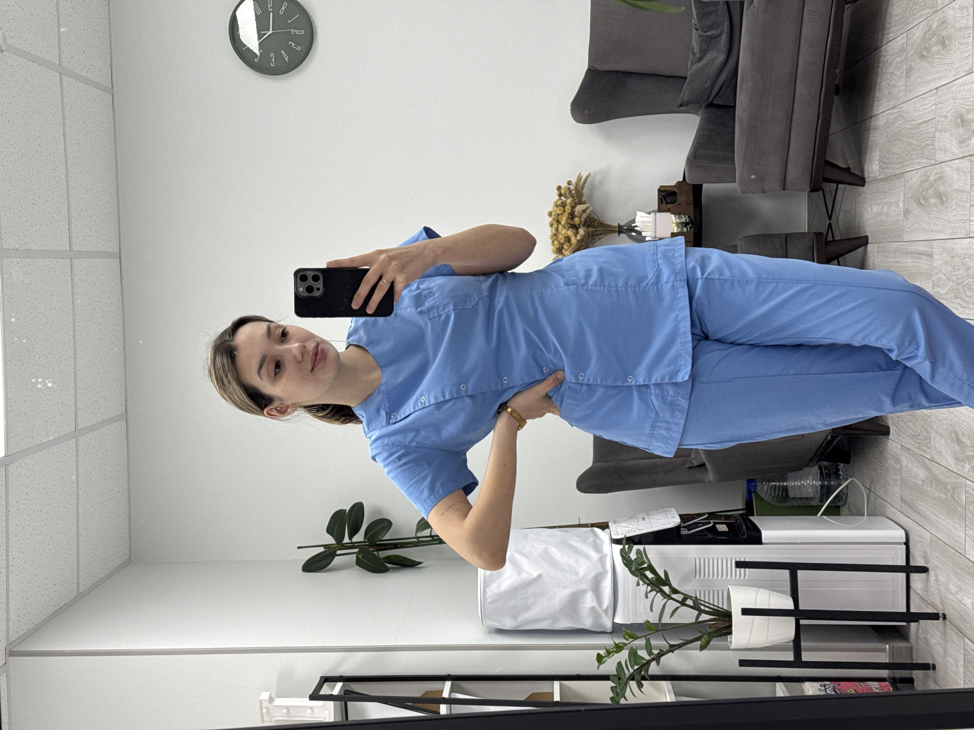 A young woman is captured in a mirror selfie, standing in what appears to be an office, clinic, or waiting room. She is looking directly at the camera, holding a black smartphone with multiple lenses up to her face with her left hand, while her right hand rests on her hip. She has light brown hair, pulled back, and is wearing a light blue short-sleeved top with buttons down the front, paired with matching light blue trousers, suggesting a medical or professional uniform. A gold-colored bracelet is visible on her right wrist, and she wears a ring on her left ring finger.
The background features white walls and a white ceiling with acoustic tiles. A round, dark green wall clock is visible in the upper right, showing the time as approximately 10:00, with numbers 12, 2, 3, 4, 8, and 10 partially visible. To the right, there are two comfortable gray armchairs or small sofas. A small table beside them holds a bouquet of dried, yellowish flowers. Below, a green potted plant sits on the floor near a low shelving unit. This unit, made of black and white elements, holds various items including a white plastic bag, what appears to be a small refrigerator or cabinet, electronic devices, a clear plastic water bottle, and another potted plant. The floor has a light wood plank design. There is no discernible text visible beyond the clock numbers. The overall atmosphere is clean and functional.