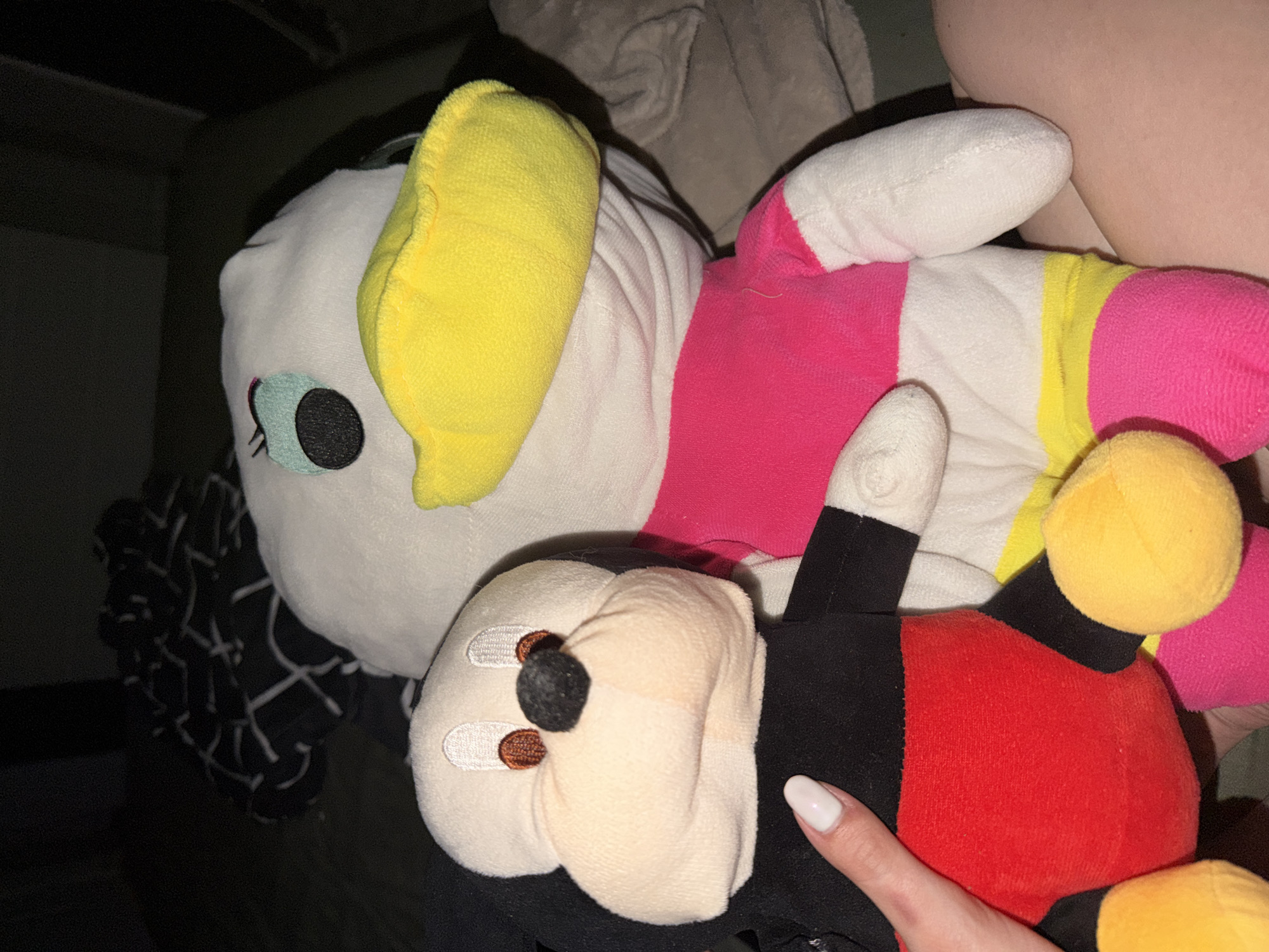 The image captures a close-up view of two plush toys, likely in an indoor, dimly lit setting. The primary subjects are a Daisy Duck plush and a Mickey Mouse plush, positioned close together, with a human hand interacting with the Mickey Mouse toy.
The Daisy Duck plush, on the left and slightly behind, is predominantly white with a large yellow bow on its head. It features a stylized teal eye with a black pupil and eyelashes on its left side. Its body is partially visible, showing sections of pink, white, and yellow fabric, suggesting a colorful outfit.
In the foreground, partially covered by the Daisy Duck plush, is a Mickey Mouse plush. It has a cream-colored face with small brown eyes and a black nose. Its body is primarily black, with a red torso and yellow feet, characteristic of the iconic Mickey Mouse outfit. A human hand, with long, oval-shaped fingernails painted white, is gently touching the Mickey Mouse plush on its black arm/side.
The background is dark and indistinct, consisting of various fabrics. To the far left, a black fabric with a subtle white linear pattern is visible. Other areas show darker, plain fabrics, possibly bedding or clothing, creating a soft, textured environment. The overall lighting is low, suggesting either nighttime or a poorly lit room, casting soft shadows. There is no visible text in the image.