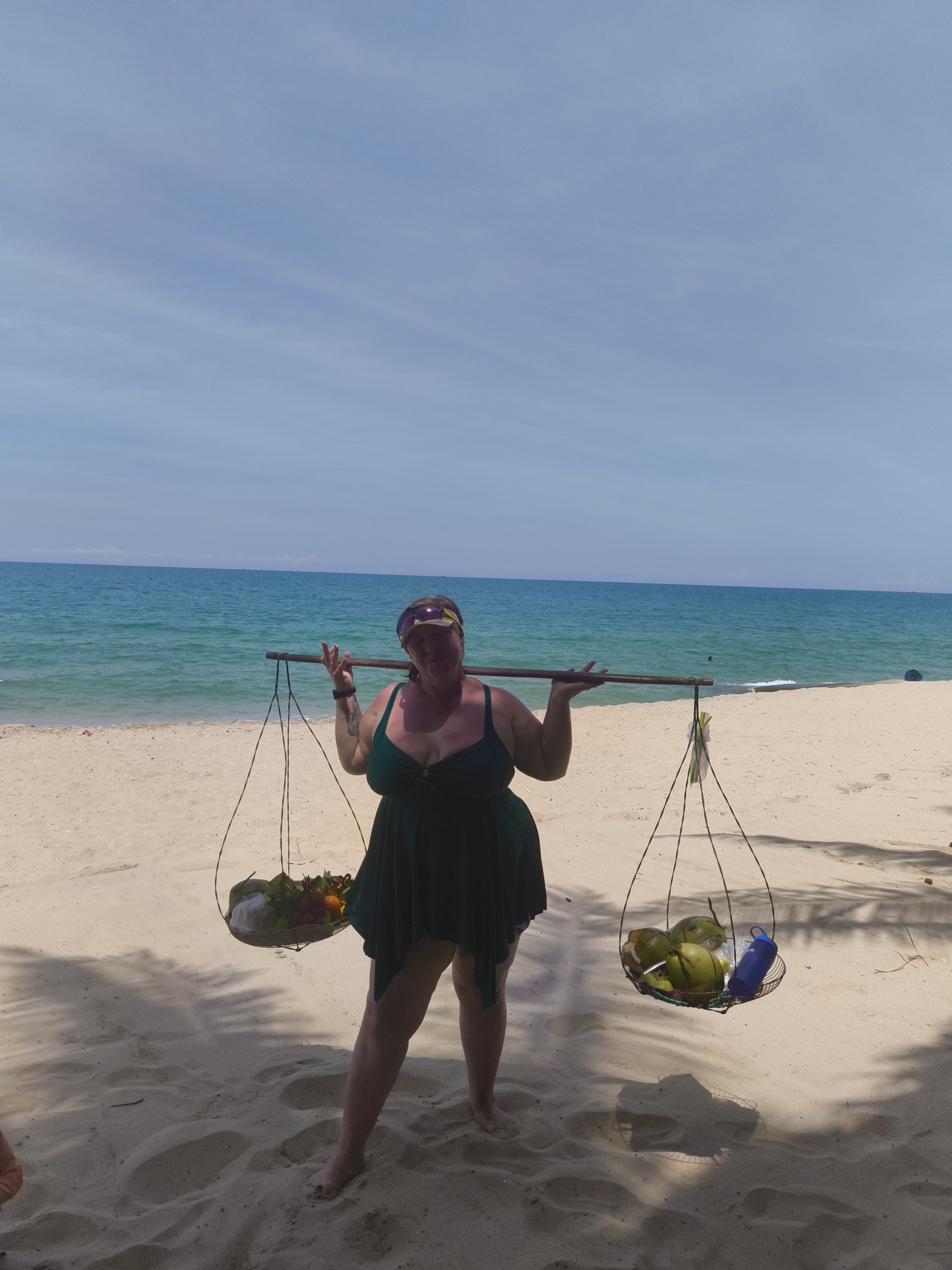 A woman stands on a sandy beach in Phu Thuy Ward, Vietnam, carrying a yoke across her shoulders. A basket hangs from each end of the yoke. The left basket contains assorted fruits, including what appear to be red and green apples, and possibly bananas. The right basket holds several green coconuts and a blue water bottle. The woman is wearing a dark green dress and a purple and yellow headband. The ocean is visible in the background, with calm turquoise water under a clear blue sky with some wispy clouds. Palm tree shadows are cast on the sand in the foreground.
