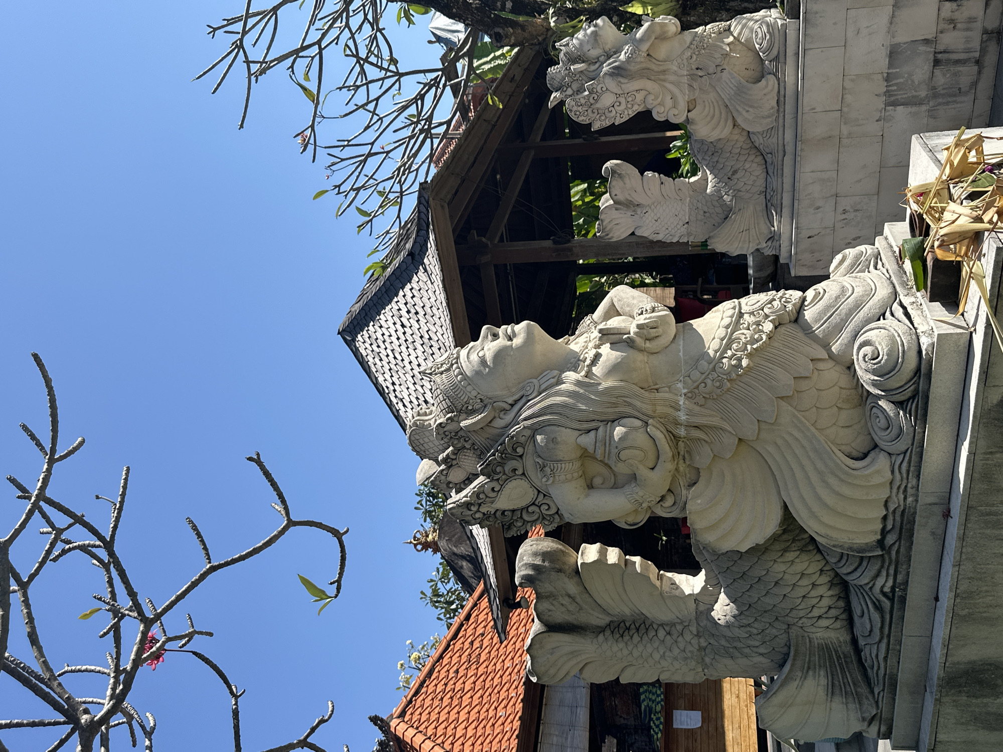 The image depicts stone sculptures of mythical figures, possibly mermaids or naga, in an outdoor setting. The sculptures are light-colored and intricately carved, with detailed scales and headdresses. They are positioned in front of traditional Indonesian architecture, characterized by red-tiled roofs and wooden structures. In the background, lush green foliage and a clear blue sky are visible, suggesting a tropical climate. The overall scene evokes a sense of cultural heritage and spirituality, likely found in a temple or historical site within Denpasar, Indonesia.