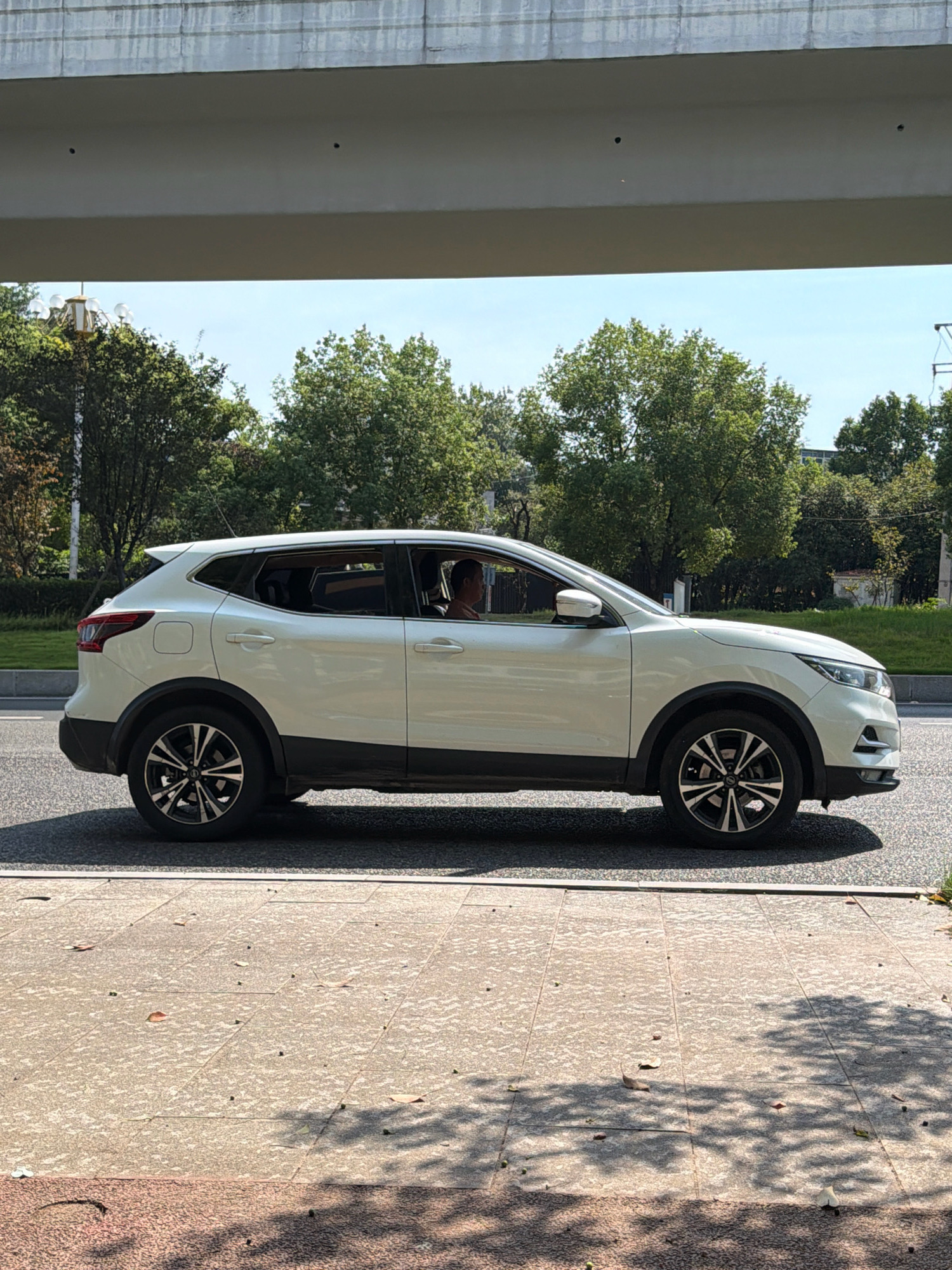 A white Nissan Qashqai is captured in a side profile view, driving on a paved road in the city of Jiangjin, China. The vehicle is positioned on the right side of the road, with its front facing towards the right of the frame. Sunlight casts shadows on the road and a light-colored tiled sidewalk. The background features lush green trees and a bright blue sky, with a concrete overpass extending overhead, obscuring a portion of the sky. A person's head is visible through the driver's side window. The scene appears to be an outdoor urban environment on a clear day.