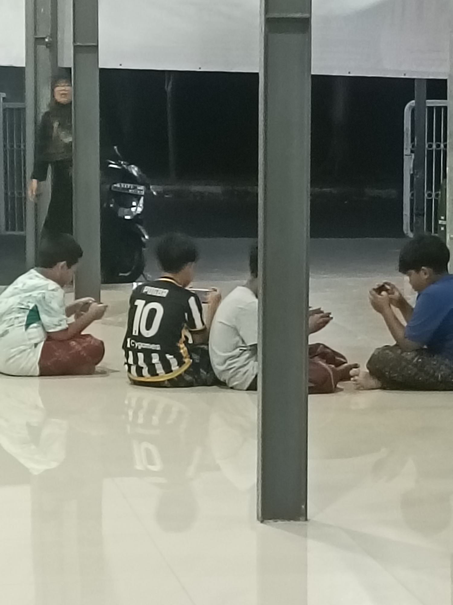 In Kediri City, Indonesia, four young boys are seated on a reflective tiled floor, engaged with their mobile devices. The scene appears to be outdoors or in a covered walkway, with tall concrete pillars supporting a roof overhead. A motorcycle is parked in the background, partially obscured by the pillars. To the left, a woman stands near a metal fence, looking towards the boys. The lighting is dim, suggesting it might be evening or night. The boys are dressed casually, with one wearing a black jersey with the number "10" prominently displayed. The floor's reflective surface mirrors the surrounding environment and the subjects within the frame.
