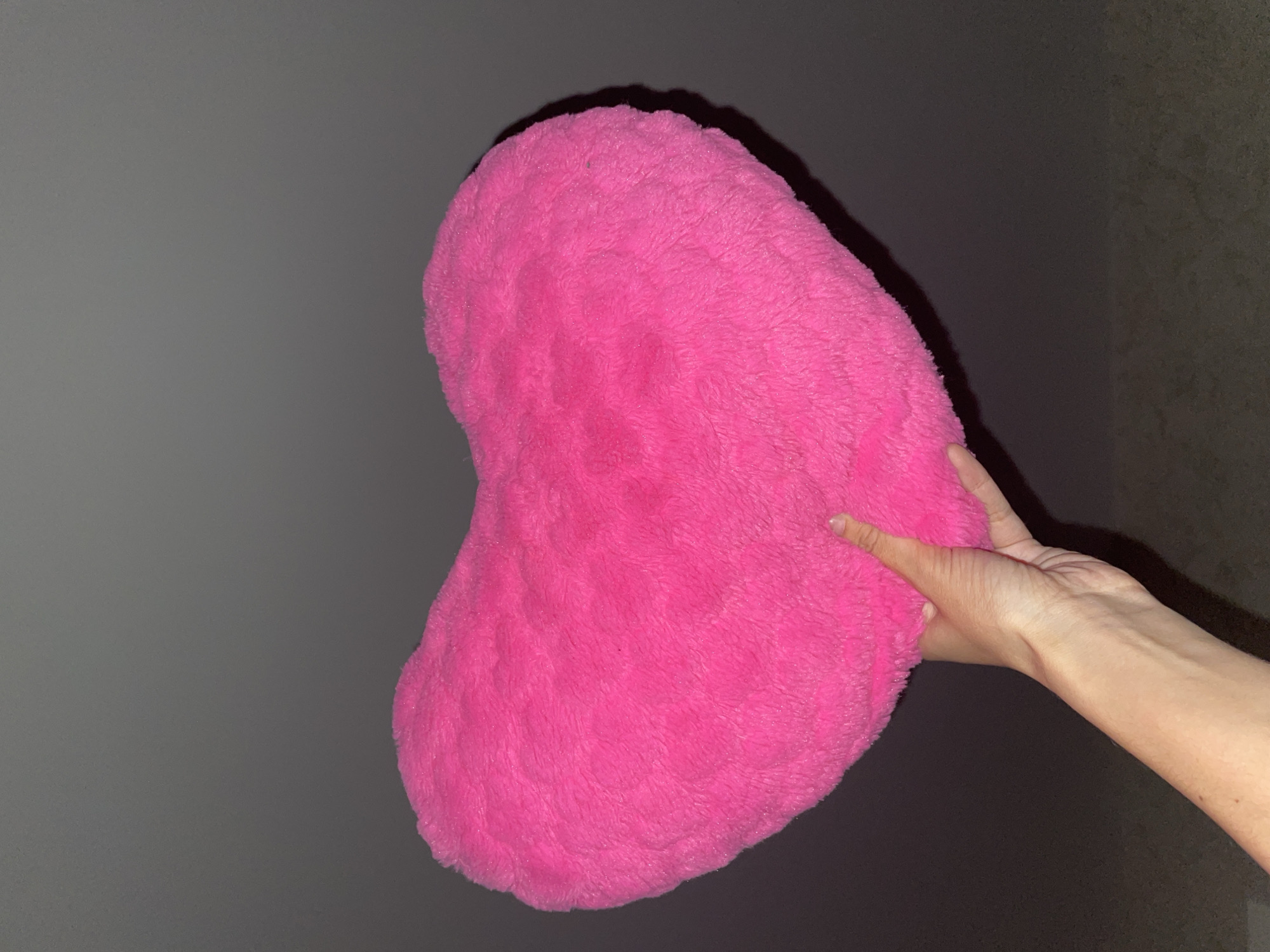A person is holding a fluffy, bright pink, heart-shaped pillow. The background is a gray wall on the left and a darker, textured wall on the right. There are no identifiable landmarks to confirm the location as Izhevsk, Russia.