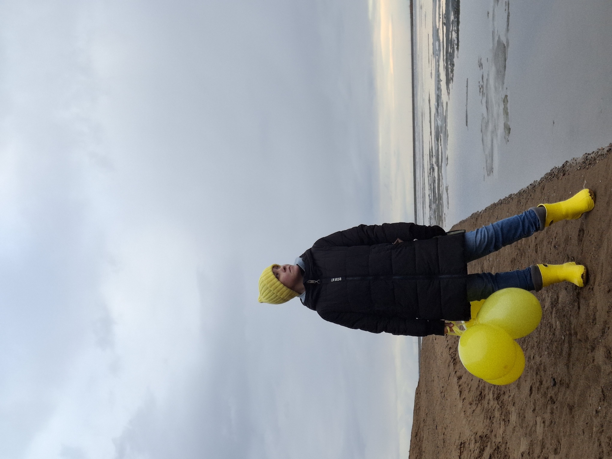 A person, appearing to be a child or young teenager, stands on a sandy or muddy shoreline next to a body of water under an overcast sky. The image is taken at a slight low angle, looking up at the subject and the vast sky.
The person is dressed for cold weather, wearing a bright yellow knitted beanie, a black puffer jacket, blue jeans, and distinctive bright yellow rain boots. They hold two vibrant yellow balloons in their right hand, which are visible near their feet. The person's gaze is directed upwards and slightly to their left, with a neutral expression. On the chest of the black puffer jacket, the text "DPEPEAT" is visible.
The setting appears to be a natural body of water, possibly a large river or lake, with some scattered ice or slush formations on the water's surface, suggesting cold temperatures. The sky is largely covered with light grey clouds, indicating an overcast day. The time of day appears to be daylight, but the subdued lighting suggests either early morning, late afternoon, or simply a heavily clouded day. The foreground consists of dark, wet sand or earth, extending to the water's edge. In the distance across the water, a faint, light-colored horizon line is visible. The overall mood is quiet and slightly melancholic, emphasized by the muted colors of the sky and water, contrasted by the bright yellow accents of the person's clothing and balloons. The location context indicates this is likely near Chaikovsky, Russia.