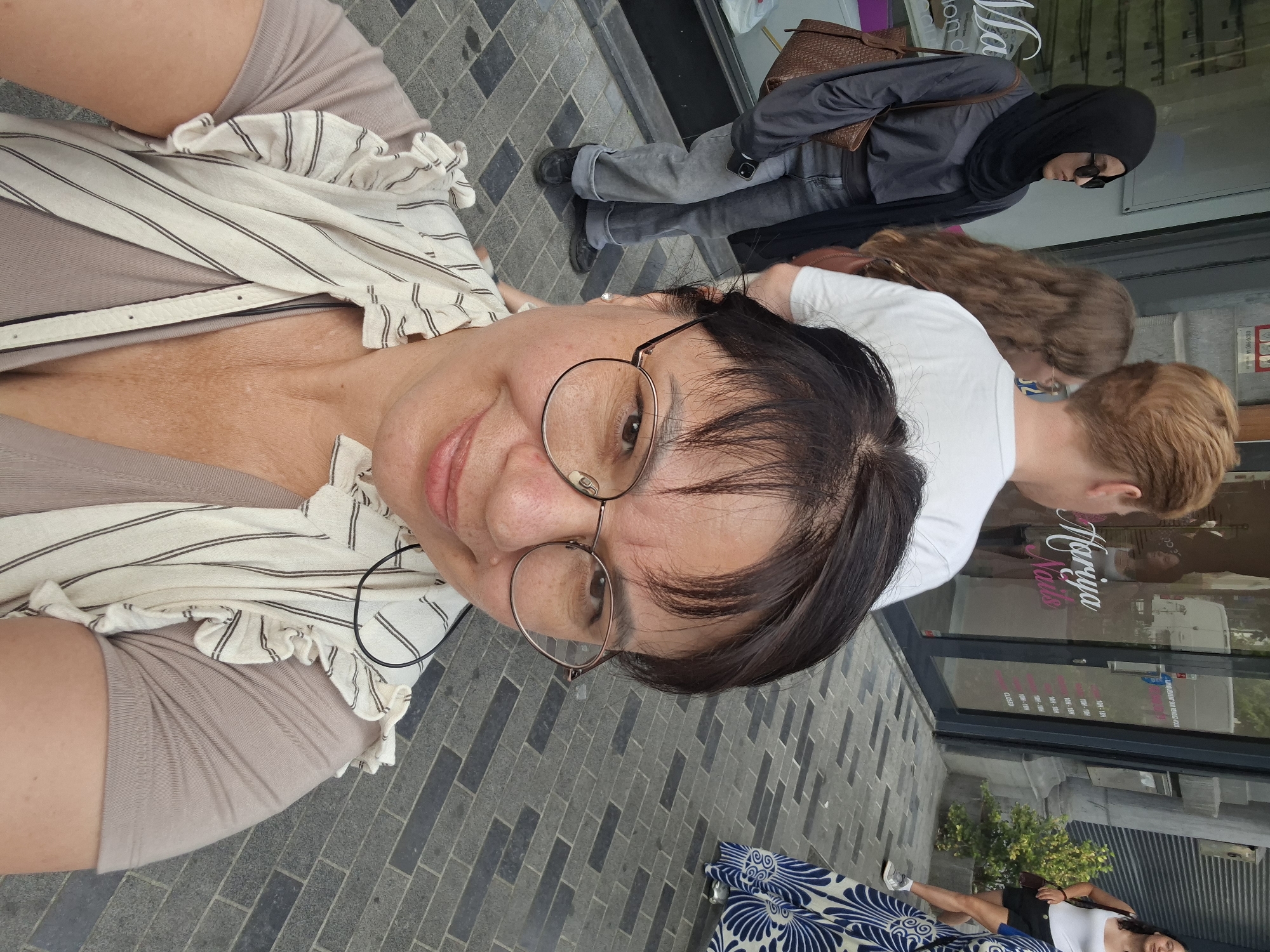 A selfie is taken from a low angle, capturing a woman in the foreground wearing glasses, a tan t-shirt, and a white and tan striped vest with ruffled sleeves. She is smiling and looking towards the camera. Behind her, several other people are visible. To her left, a woman wearing a black hijab and sunglasses stands with a brown purse over her shoulder, looking to her left. In the middle ground, a person with reddish-blonde hair wearing a white t-shirt is walking away from the camera. To the right, a woman in a white tank top and black shorts is walking away from the camera. The background shows a storefront with the name "Hariya Nails" and opening hours displayed on the glass door. The ground is paved with grey rectangular bricks. The overall scene appears to be a street in a city, likely Brussels, Belgium, given the presence of the "Hariya Nails" store, a common type of establishment in urban areas.