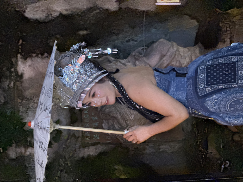 A female subject of East Asian appearance is captured from the mid-torso upward, positioned against a textured stone wall in what is identified as Yuzhong District, China. She gazes to her right with a slight smile. Her attire includes an elaborate, silver-toned headpiece featuring reflective surfaces, a red-orange decorative element on the upper right, and several dangling, multi-segment ornaments on the right side. Glitter, possibly shaped like butterflies, is applied below her eyes. She wears a strapless blue garment characterized by bandana-style patterns and a structured silhouette. Her right hand, adorned with a silver ring on the ring finger, holds a white paper umbrella featuring dark, possibly calligraphic or floral, patterns. The umbrella has a light-colored wooden shaft and a red finial. The background consists of a dark, uneven rock or stone wall, with Chinese characters carved into it, notably '大' (dà), visible behind the umbrella handle. The scene is illuminated by dim, directed lighting.