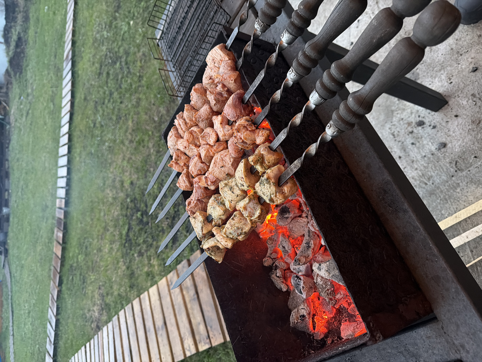 The image shows a barbecue grill with several skewers of meat cooking over hot coals. The skewers have twisted metal shafts and wooden handles. The meat on the skewers varies in color and appearance, suggesting different marinades or types of meat. The grill is set up outdoors, with grass and a decorative border visible in the background. The location is identified as Большая Шильна, Russia.