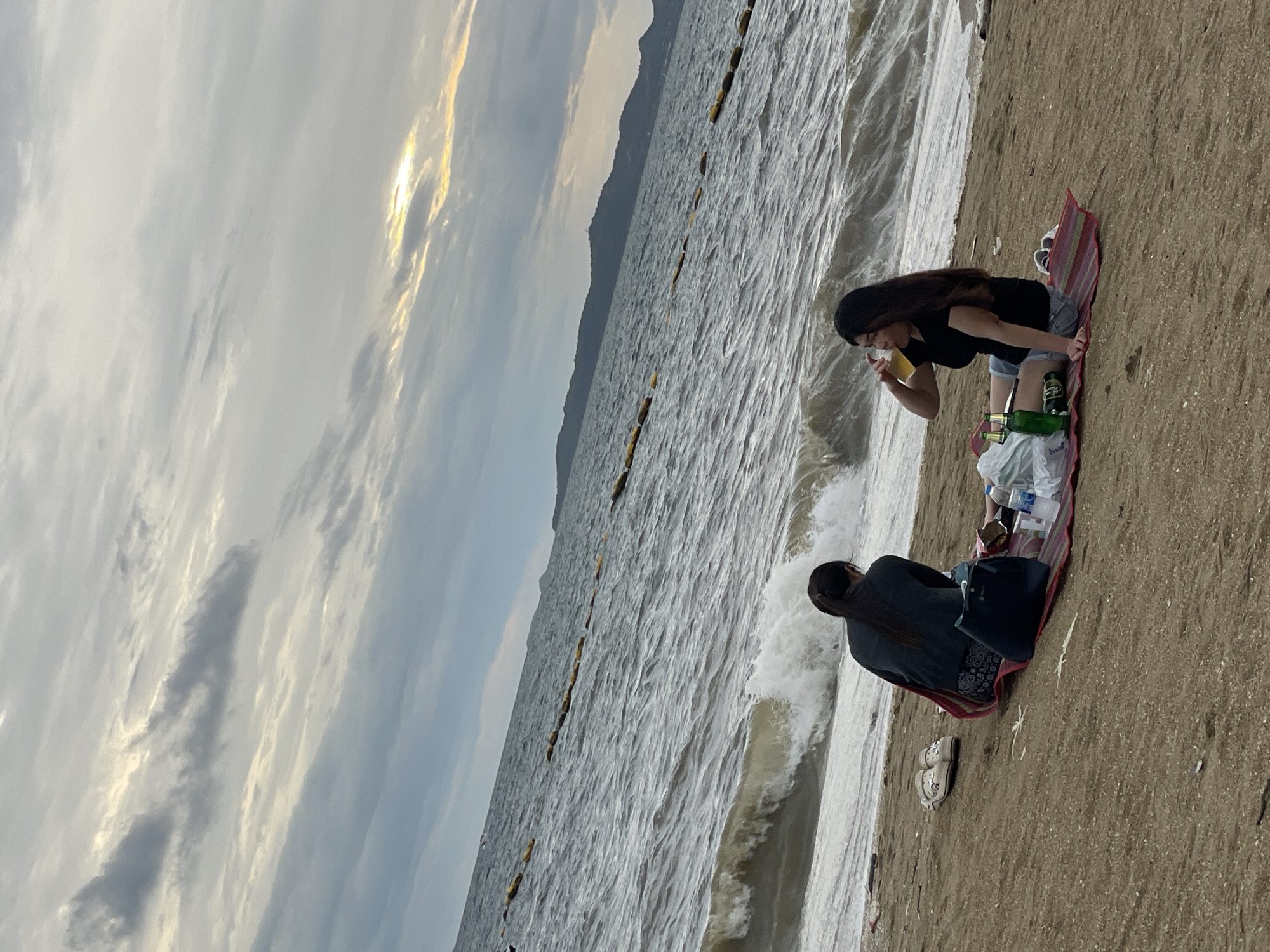 Two women are having a picnic on Jomtien Beach in Pattaya City, Thailand. The woman on the right is wearing a black t-shirt and denim shorts, and is drinking from a glass of beer. The woman on the left is wearing a dark jacket and patterned skirt, and is looking out at the ocean. They are sitting on a mat on the sand, with a cooler bag, bottles of beer, and other picnic items spread around them. The ocean is rough, with large waves crashing on the shore. The sky is cloudy, with some patches of sunlight breaking through. In the background, there are hills and a breakwater.