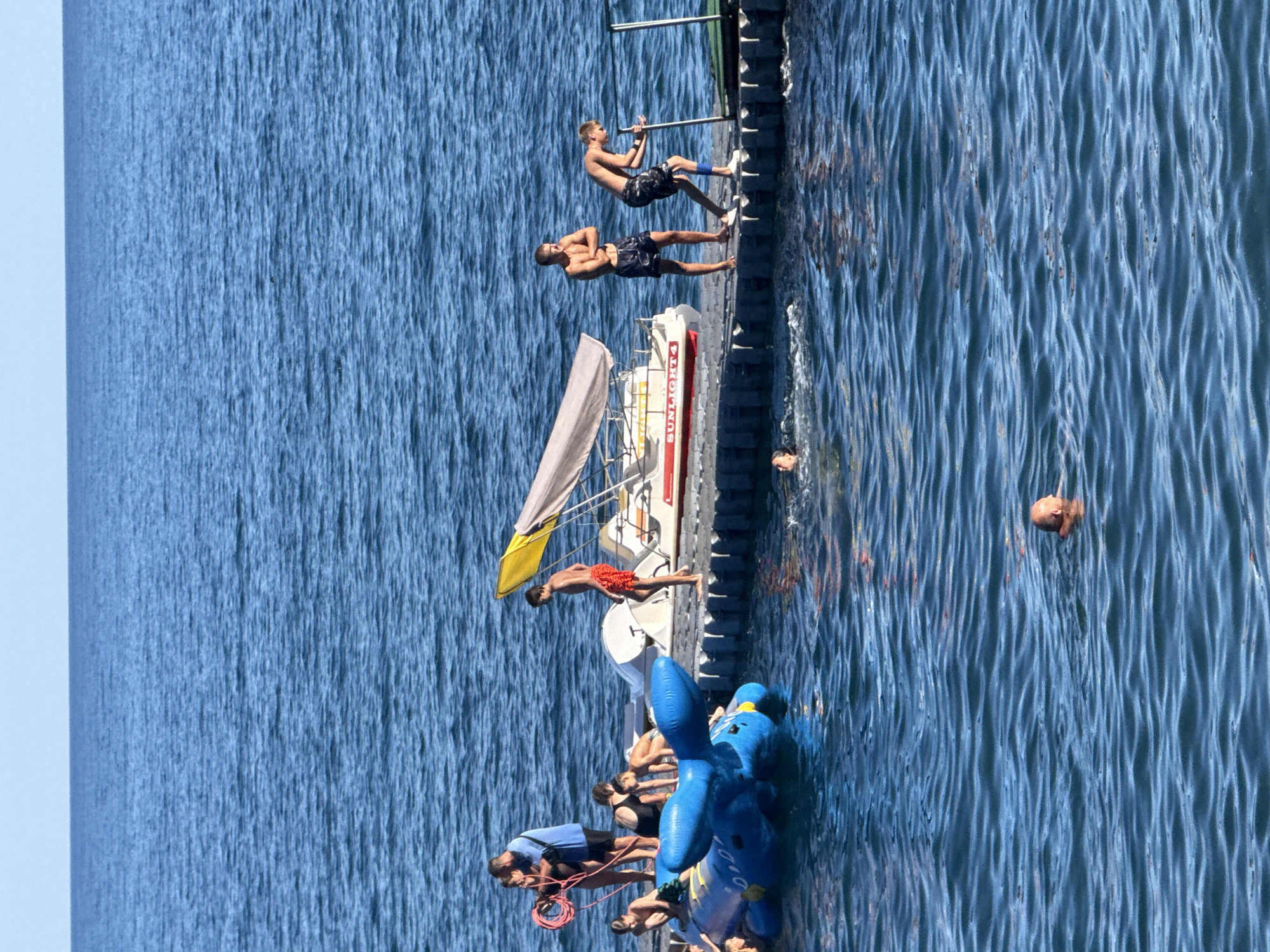 The image depicts a scene at the seaside in Partenit, Ukraine, on a clear, sunny day. A group of people are gathered on a floating platform or dock, engaged in recreational activities. Several individuals are in swim trunks and appear to be enjoying the water. One person is on the platform, holding a red coiled rope, possibly preparing for an activity or securing something. Another person is near a yellow and white canopy boat. A blue inflatable toy, resembling a whale or dolphin, is floating in the water near the dock, with a few people interacting with it. Two men are standing on the dock, one with his arms crossed and the other leaning on a pole or handle. In the water, a man with a bald head is visible swimming. The sea is calm with gentle ripples, and the sky is a clear blue. The overall atmosphere suggests a day of leisure and summer fun.