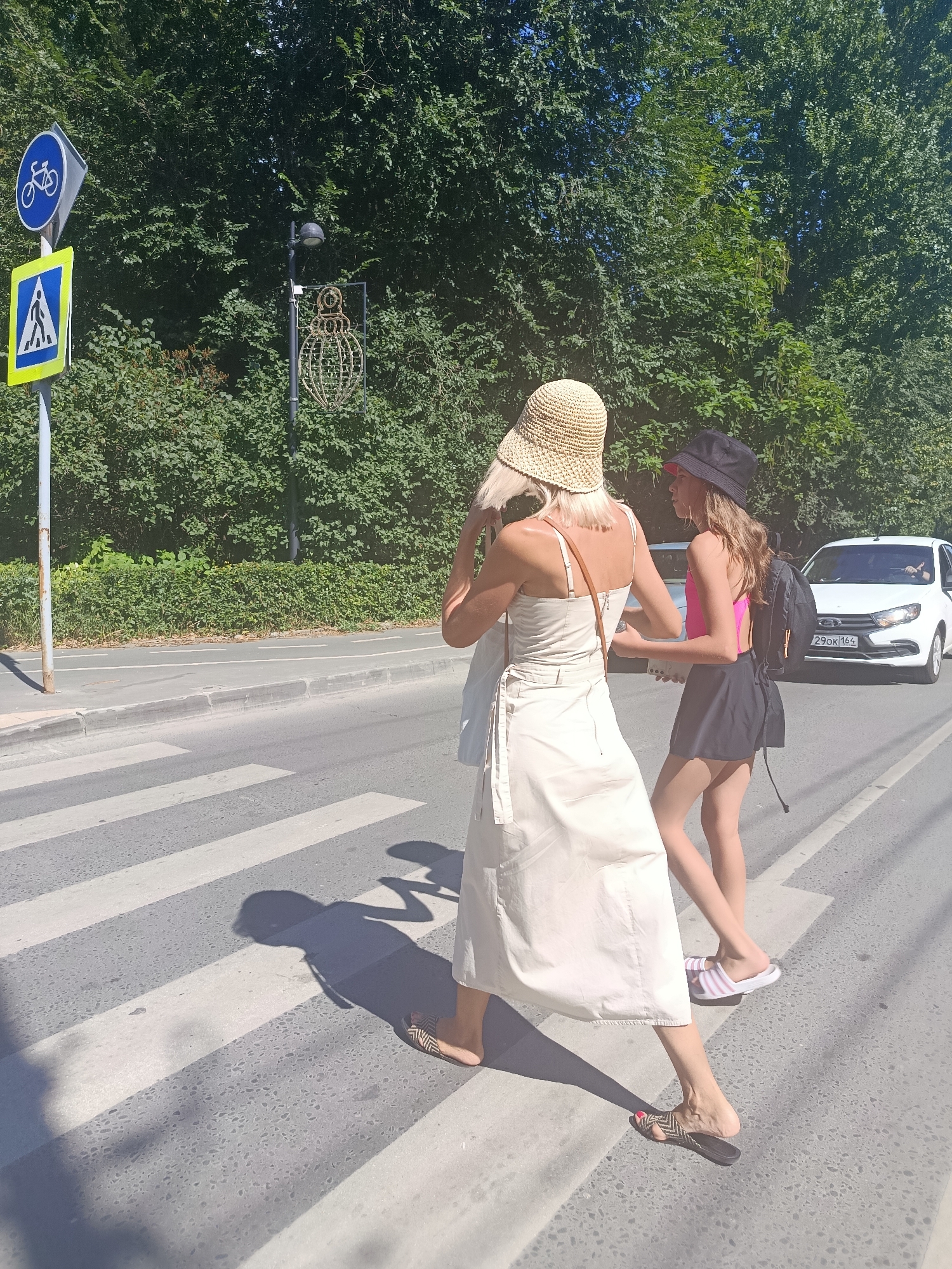 Two individuals, appearing to be women, are crossing a pedestrian walkway on a sunny day. The person in the foreground is wearing a light-colored, sleeveless dress and a woven hat. The second person, slightly behind and to the right, is wearing a pink top, a dark skirt, and a black hat. They are both wearing sandals. A white car with a license plate that reads "290OK 164" is approaching from the right. In the background, there is a densely wooded area. To the left of the frame, a blue circular sign with a bicycle symbol and a yellow square sign with a pedestrian symbol are visible on a pole, indicating a bike lane and a pedestrian crossing. The overall environment suggests an urban street scene in Saratov, Russia.