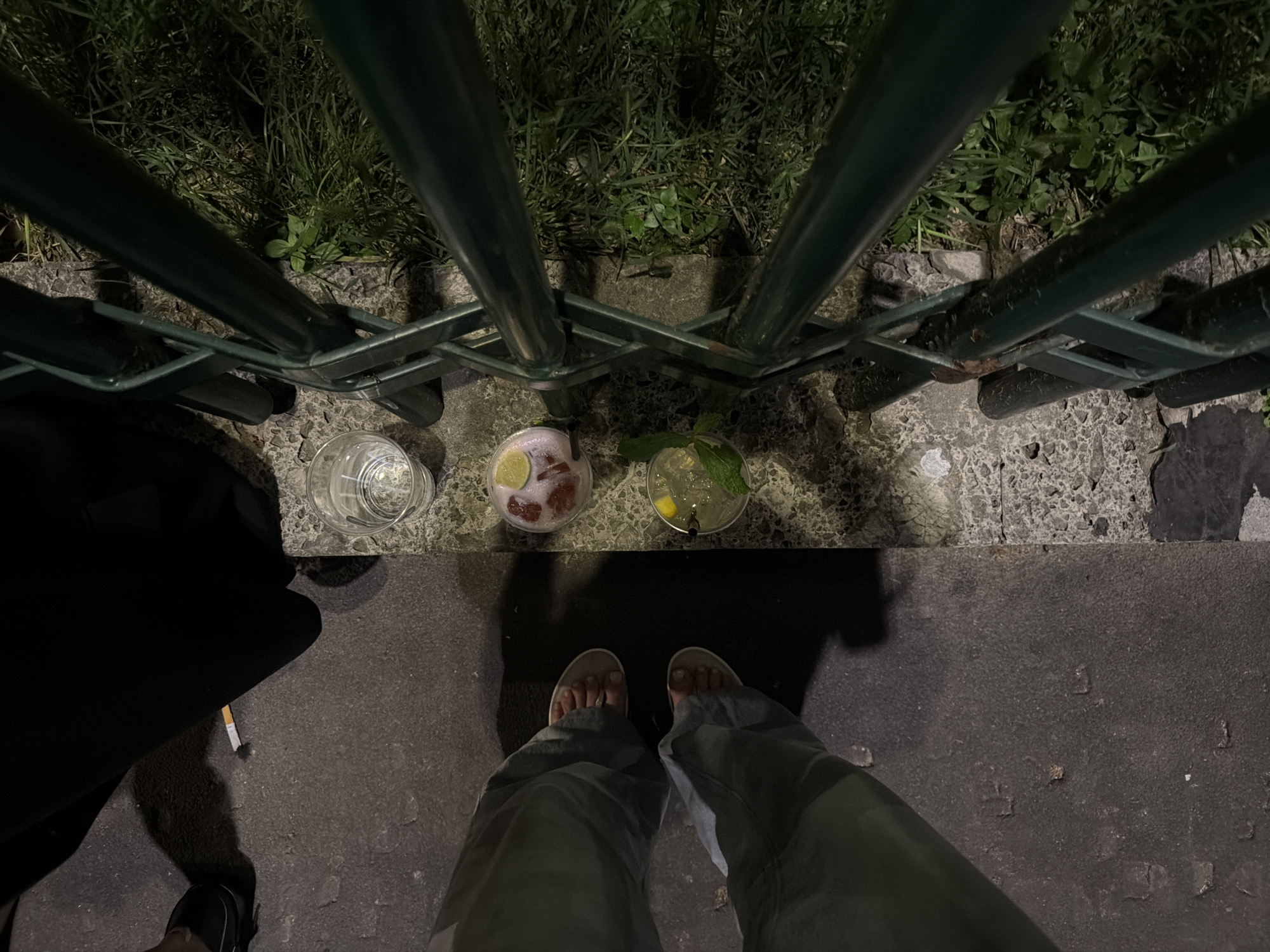 A high-angle shot shows a scene on a street or outdoor patio, likely at night, with three beverages in clear glasses sitting on the ground near a low, dark green metal fence. The first drink appears to be water, the second a pink-red drink with a lime wedge, and the third a lighter colored drink with mint sprigs. Below the drinks are a pair of feet wearing sandals. To the left, part of another person’s leg is visible, wearing a black shoe. A lit cigarette butt sits near the shoe. The setting appears to be an urban area with a rough stone ground surface and grass and greenery behind the fence. There is no way to confirm the exact location in Milan, Italy based on the visual data provided.