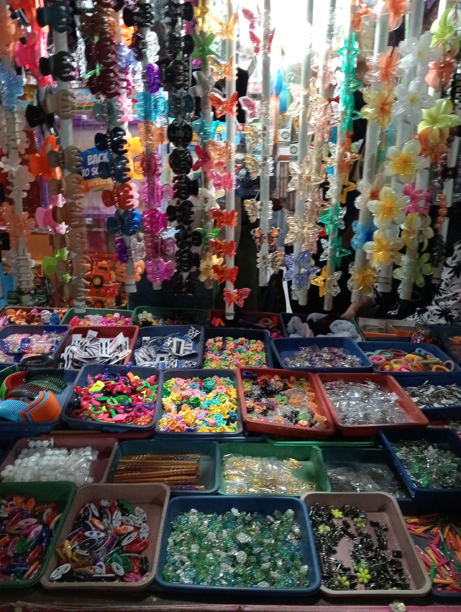 The image displays a market stall in Grabag, Indonesia, filled with various hair accessories. The stall is densely packed with colorful items, primarily hair clips and bands, arranged in numerous plastic trays and hanging displays. The lighting is bright, suggesting an indoor or covered market setting. The sheer volume of merchandise suggests a retail environment focused on affordable accessories. No people are clearly visible, but the presence of a stall indicates commercial activity. The overall impression is one of abundance and variety, typical of a bustling local market.