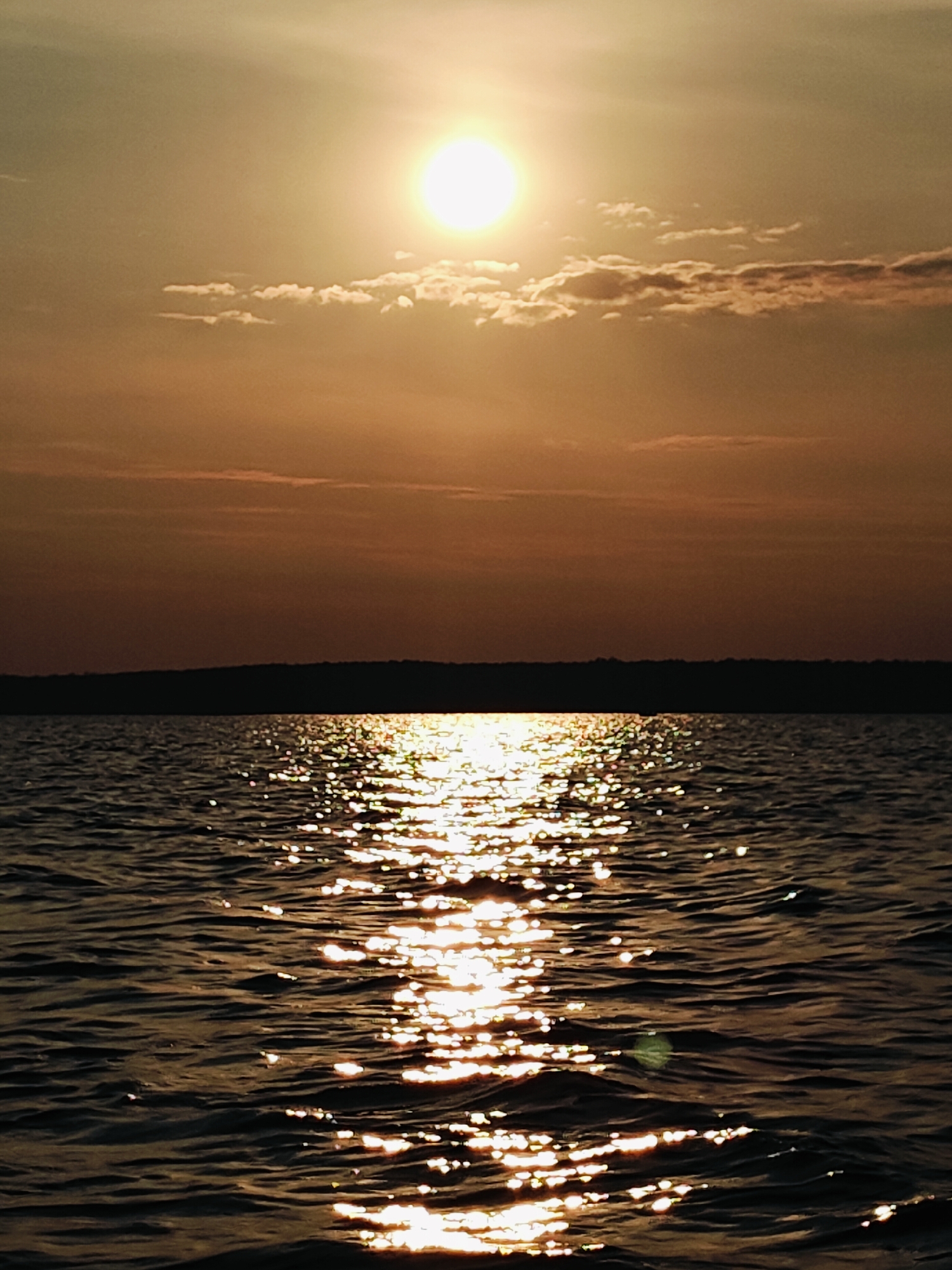 The image shows a body of water, likely a lake or river, with the sun setting in the distance. The sun is bright and positioned in the upper center of the frame, casting a warm, golden glow across the scene. Its light reflects off the rippling water, creating a shimmering path of light that extends towards the foreground. The horizon is defined by a dark, silhouetted landmass, possibly a shoreline with trees. The sky above transitions from a brighter, sunlit area near the horizon to a darker, more muted tone towards the top of the image, with a few scattered clouds. The overall impression is one of a serene and tranquil natural landscape at dusk. The image is not possible to locate in Chaykovsky, Russia without further contextual information.