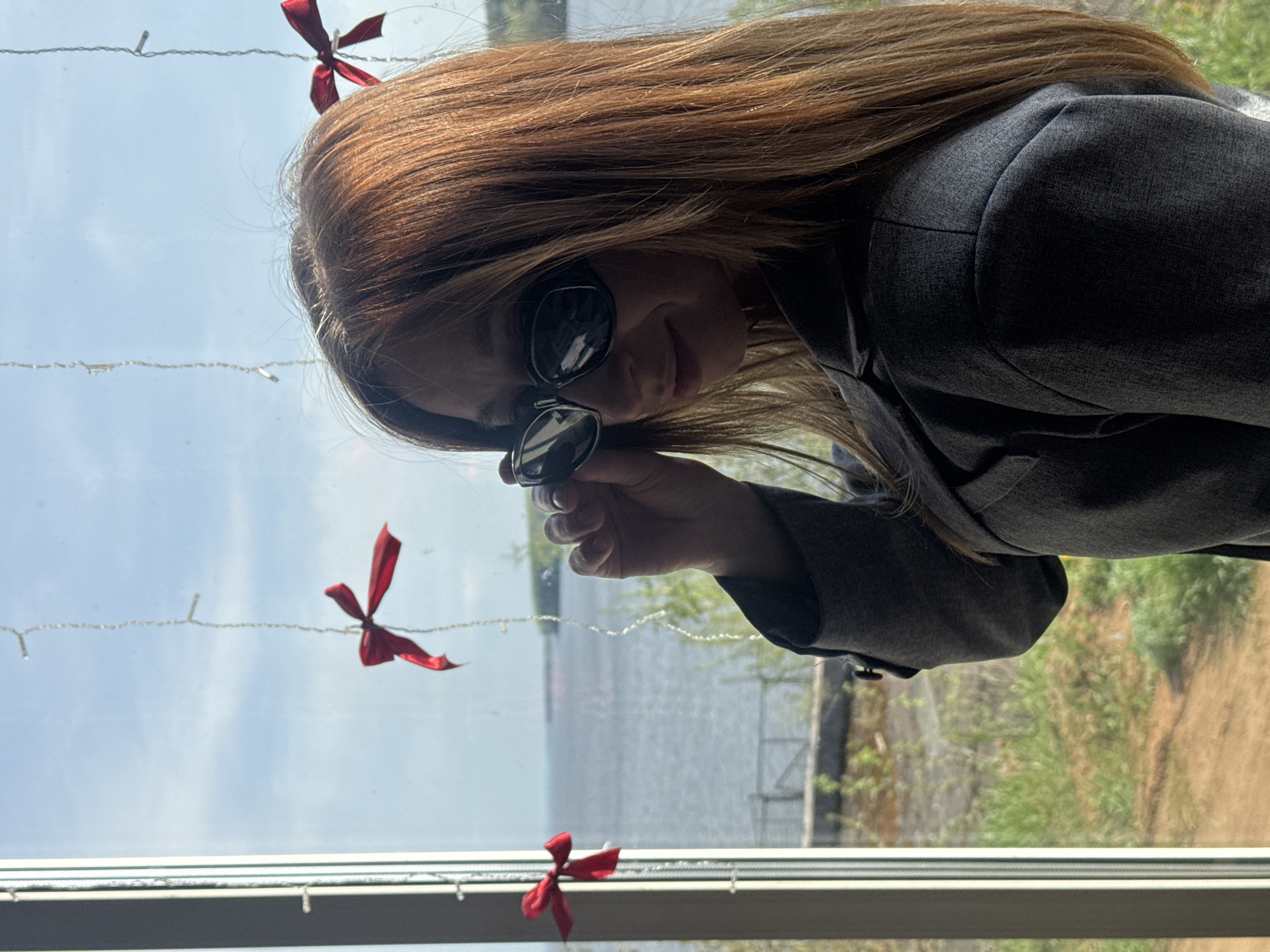 A person with long, reddish-brown hair is partially visible, wearing a gray blazer and sunglasses. They are holding the sunglasses slightly down, peering over them. In the background, a window looks out onto a body of water. Red ribbon decorations are hung on the window frame. Outside the window, there's greenery and what appears to be a shoreline. The location is identifiable as Chaikovsky, Russia, due to the specific lake features and embankment details visible outside the window.