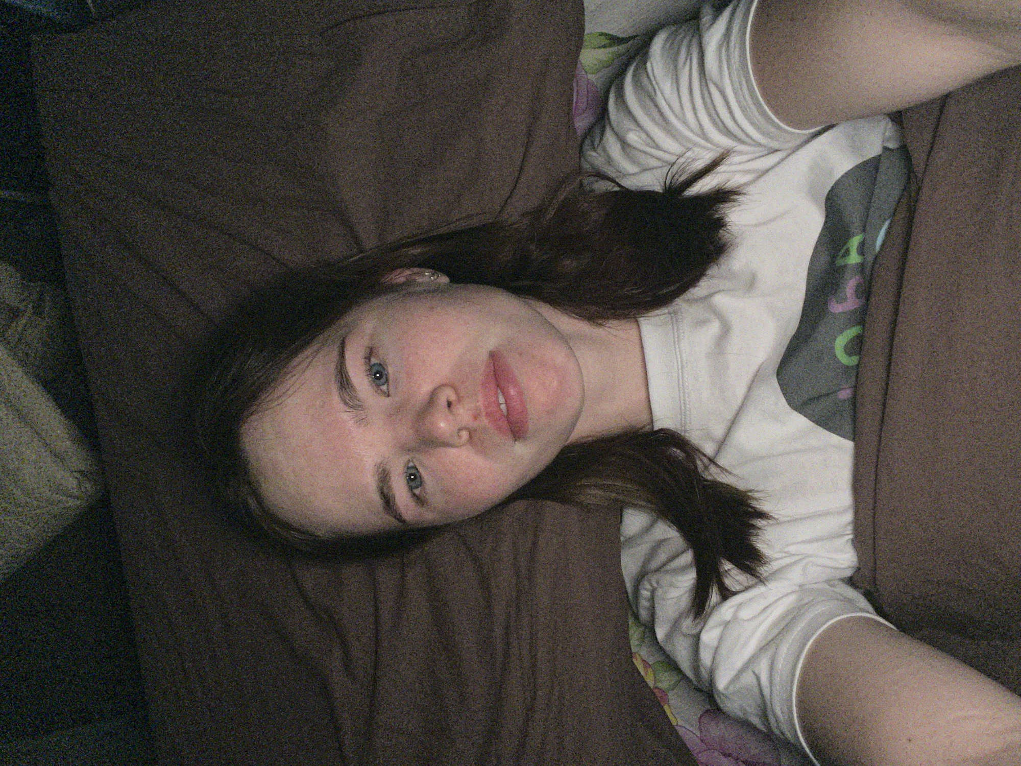 A young adult female is depicted in a close-up, head-and-shoulders selfie, looking directly at the camera. She has fair skin, light blue eyes, and long, dark blonde or light brown hair parted centrally. Her lips appear full. She is wearing a white, long-sleeved shirt with a partially visible gray graphic featuring green and pink elements on the chest. A small silver earring is visible in her left earlobe. She is positioned against and partially covered by a plain brown blanket or duvet, which extends into the background. A section of multi-colored patterned fabric, exhibiting pink, green, and purple hues, is visible to the left, consistent with additional bedding. The lighting is dim, and the image displays noticeable digital noise. The specific location within the city of Chaikovsky, Russia, cannot be determined from the visual information provided in the image.
