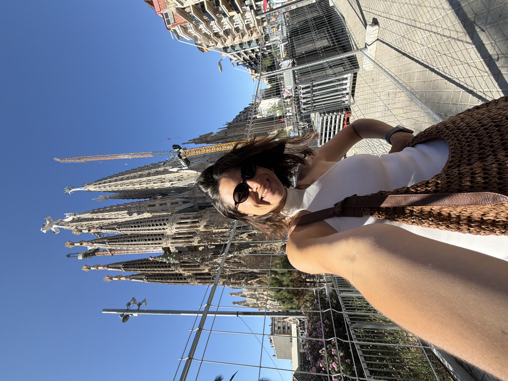 A woman with dark hair, wearing sunglasses and a white tank top, takes a selfie outdoors. She is holding a woven brown bag. Behind her, the Sagrada Familia, a large basilica in Barcelona, Spain, is visible under a clear blue sky. Construction cranes are present near the basilica, indicating ongoing work. A metal fence separates the woman from the basilica and the surrounding area, which includes trees and buildings. The ground is paved with square tiles, and shadows are cast by the sun.