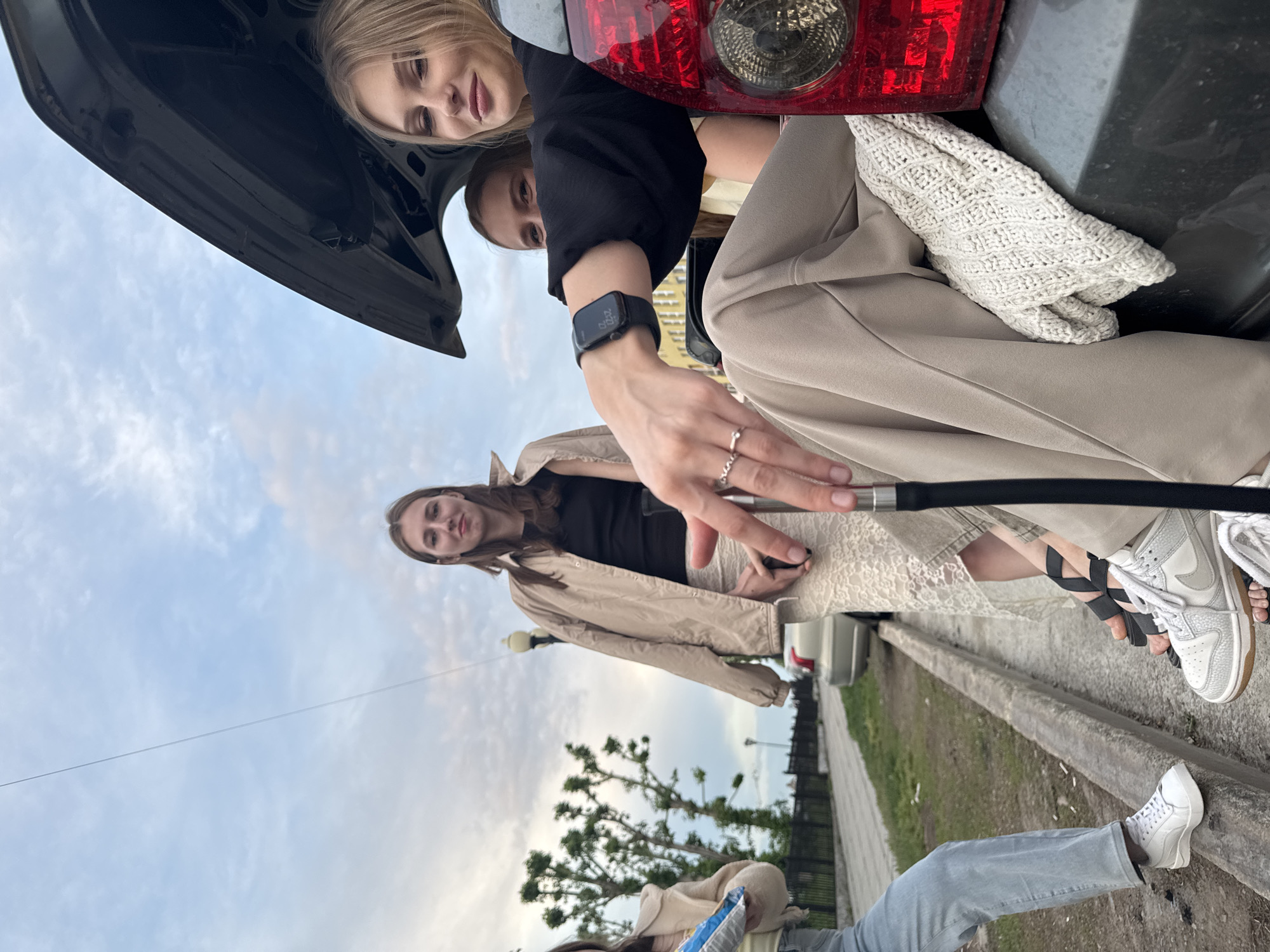 Three young women are interacting with a car in an urban environment. Two of the women are positioned in the open trunk of a gray vehicle. The trunk is open, with the car's tail light visible. One woman inside the trunk is blonde and wearing a black top. The other woman has dark hair and is looking up. A third woman stands outside the car, dressed in a tan jacket over a black top, and beige pants. She is holding a black object that extends towards the trunk. All are white. The surrounding environment includes a curb, pavement, a metal fence, grass, and trees. A street lamp is visible in the background against a cloudy sky. The location is Perm, Russia.