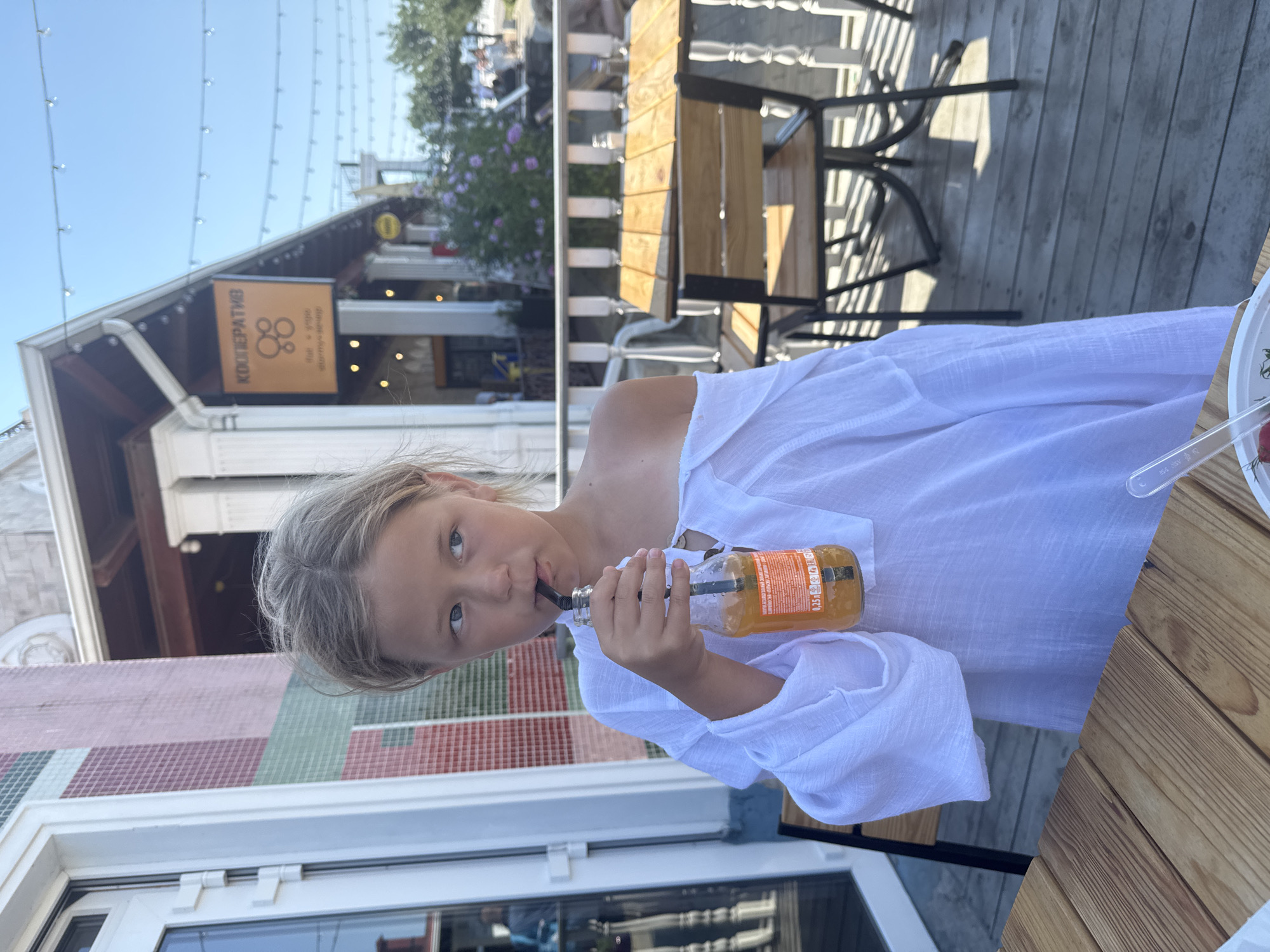 A young girl with blonde hair is seated outdoors at a wooden table, drinking from a small glass bottle of orange-colored liquid. She wears a white, off-the-shoulder top. The setting appears to be a cafe or restaurant with a wooden deck and outdoor seating, including metal chairs and tables with wooden slats. In the background, there is a building with a sign that has Cyrillic text and a circular logo, along with some greenery and a clear sky. The time of day appears to be daytime, with bright sunlight casting shadows. The girl's expression is relaxed as she sips from the bottle with a straw. Visible text on the bottle includes "0,25 л" and other unreadable Russian characters. The location context is Gelendzhik, Russia.