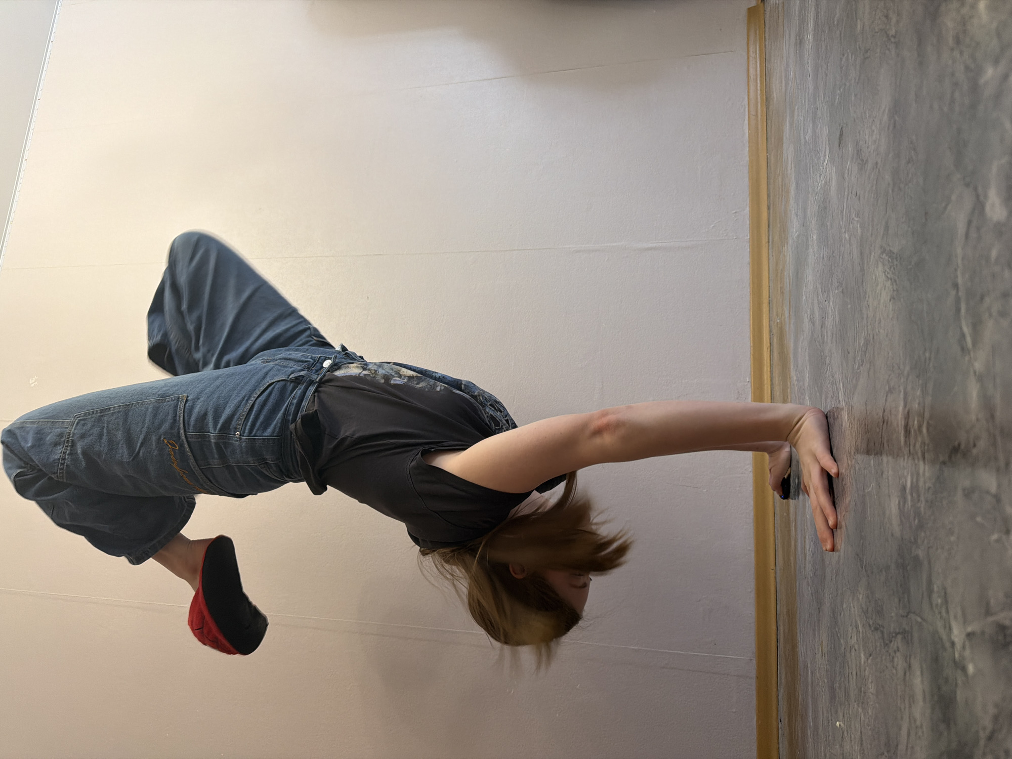 The media file displays a person captured in an acrobatic or dance-like pose within an indoor setting. The image is oriented sideways, with the person positioned horizontally. The subject, a young individual, wears a black t-shirt and loose blue denim jeans, which feature faint embroidery on the back pocket, possibly reading "Desigual." Their medium-length brown hair is fanned out, indicating movement or the effect of gravity given the unusual orientation. A red and black slipper or sock-like footwear is visible on the left foot.
The background consists of two distinct surfaces: a plain, light beige or off-white wall that covers most of the frame, and a darker, greyish, textured wall or panel on the right, separated by a yellowish-gold trim. The person's right hand is pressed flat against this darker, textured surface, providing support for their body, which is suspended parallel to the lighter wall.
The activity appears to be a demonstration of physical skill, balance, and strength, possibly part of a training session or performance. The pose involves the person's body being held almost entirely off the ground (relative to a standard vertical orientation of the walls). The setting is likely a simple room or studio, with artificial lighting providing even illumination. No specific time of day or emotions are clearly discernible. The location context provided is Perm, Russia.