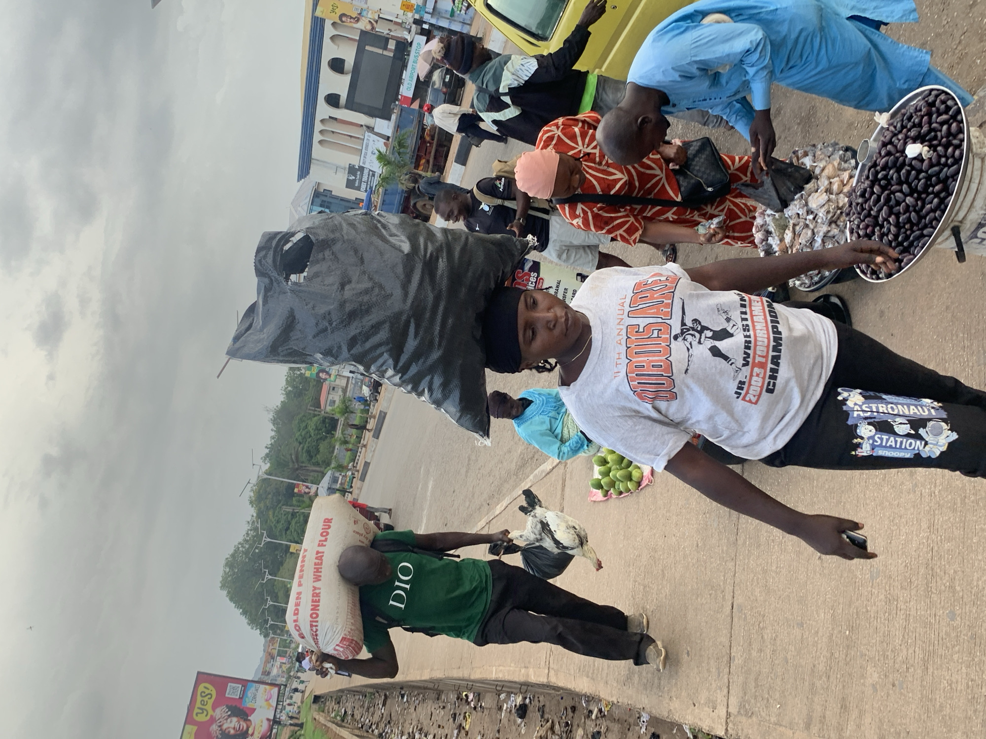 In Barakin Lerawi, Nigeria, a bustling street scene unfolds under an overcast sky. A man in a green t-shirt walks along a sidewalk, balancing a large sack of "GOLDEN PENNY CONFECTIONERY WHEAT FLOUR" on his head and carrying a fowl in a plastic bag. He is dressed in black trousers and beige shoes. Beside him, a woman wearing a black headscarf and a white t-shirt with graphics walks towards the camera, holding a mobile phone in her left hand. Behind her, a vendor in a red and white patterned dress is seen. A man in a bright blue outfit is interacting with a stall displaying small, shelled items in a large bucket and a tray filled with dark, oval fruits, possibly olives. Another man in a blue shirt and patterned hat stands near a yellow vehicle. The environment shows a street with traffic, buildings, and streetlights, indicating an urban setting with commercial activity.