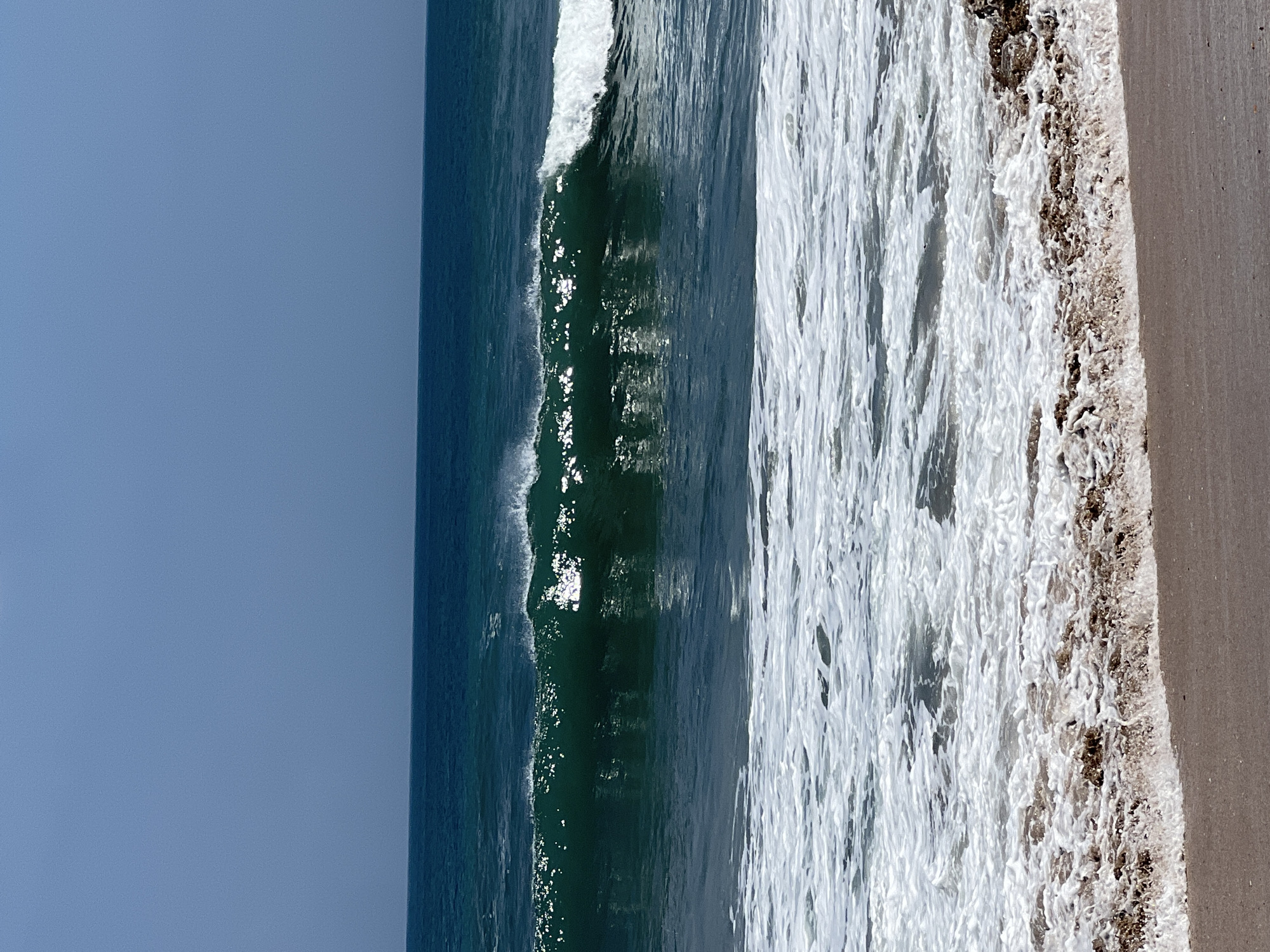 An ocean wave is breaking in the foreground of this image, with white foam washing onto a sandy beach. The water is a deep blue-green, and the sky is a clear, pale blue. The scene is tranquil and serene, evoking a sense of peace and relaxation. The image appears to have been taken on a sunny day, with the sun reflecting off the water. It is a beautiful depiction of a tropical beach.