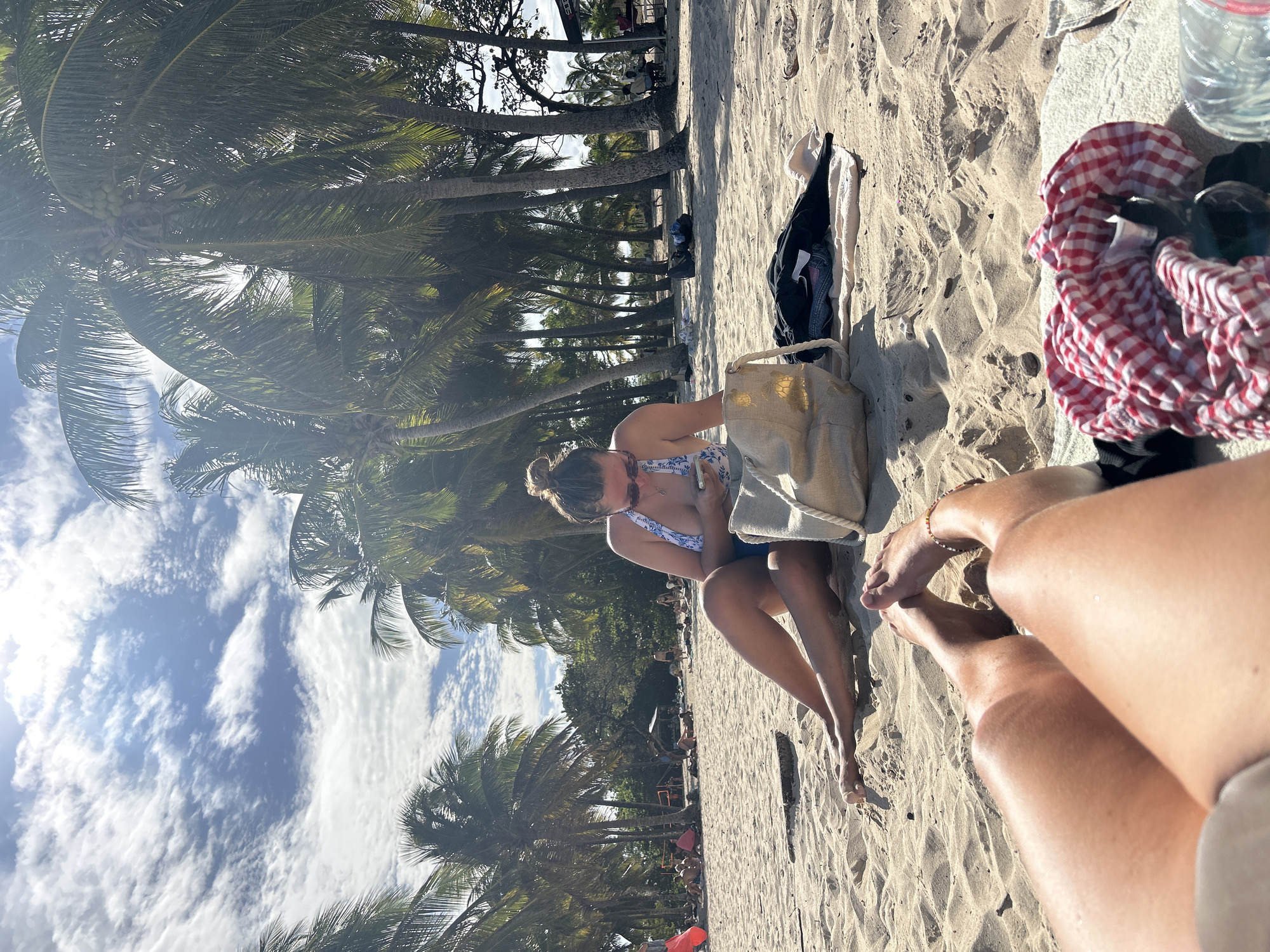 On a beach in Sainte-Anne, France, a woman sits on the sand near palm trees, looking at her phone while leaning against a beige bag. A towel with a red and white checkered pattern is nearby, along with other bags. Another person's legs and feet are visible in the foreground. The sky is partly cloudy.