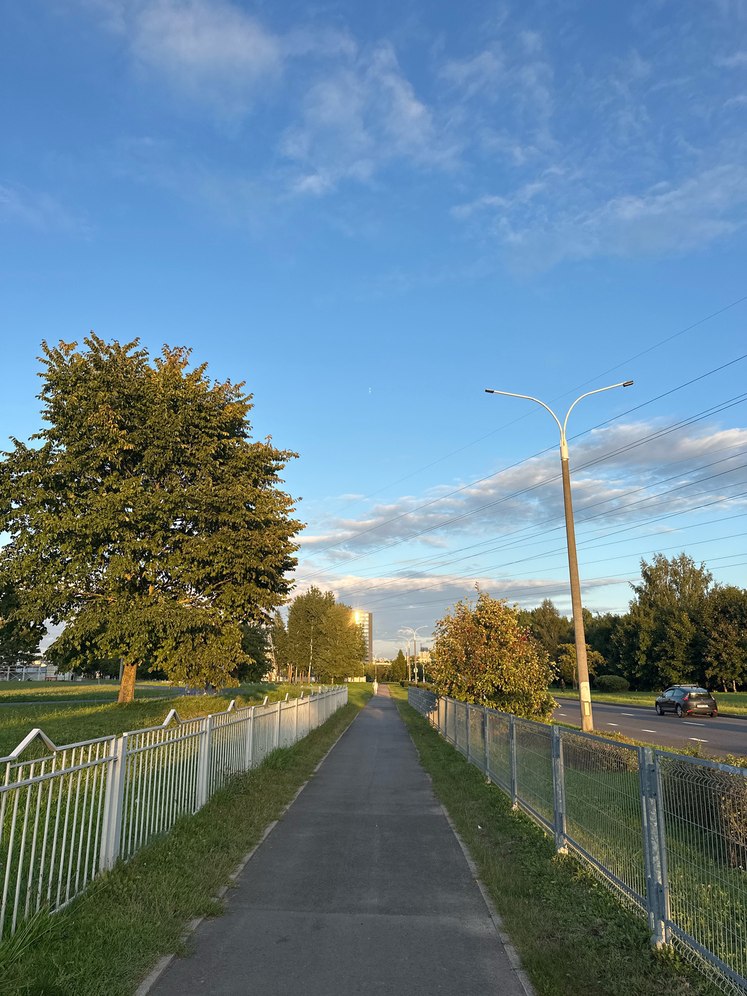 A paved pedestrian path runs diagonally from the bottom center towards the horizon in a city setting. The path is bordered on the left by a white metal fence and green grass, and on the right by a darker metal fence and more grass. Beyond the fences, trees and a road with a dark-colored car are visible. A tall street light with two lamps stands near the right side of the path. The sky is blue with scattered white clouds, and the setting sun casts a warm glow on some of the foliage and buildings in the distance. A building resembling a modern high-rise is visible on the horizon. The scene appears to be a public park or walkway in Minsk, Belarus, during the evening.