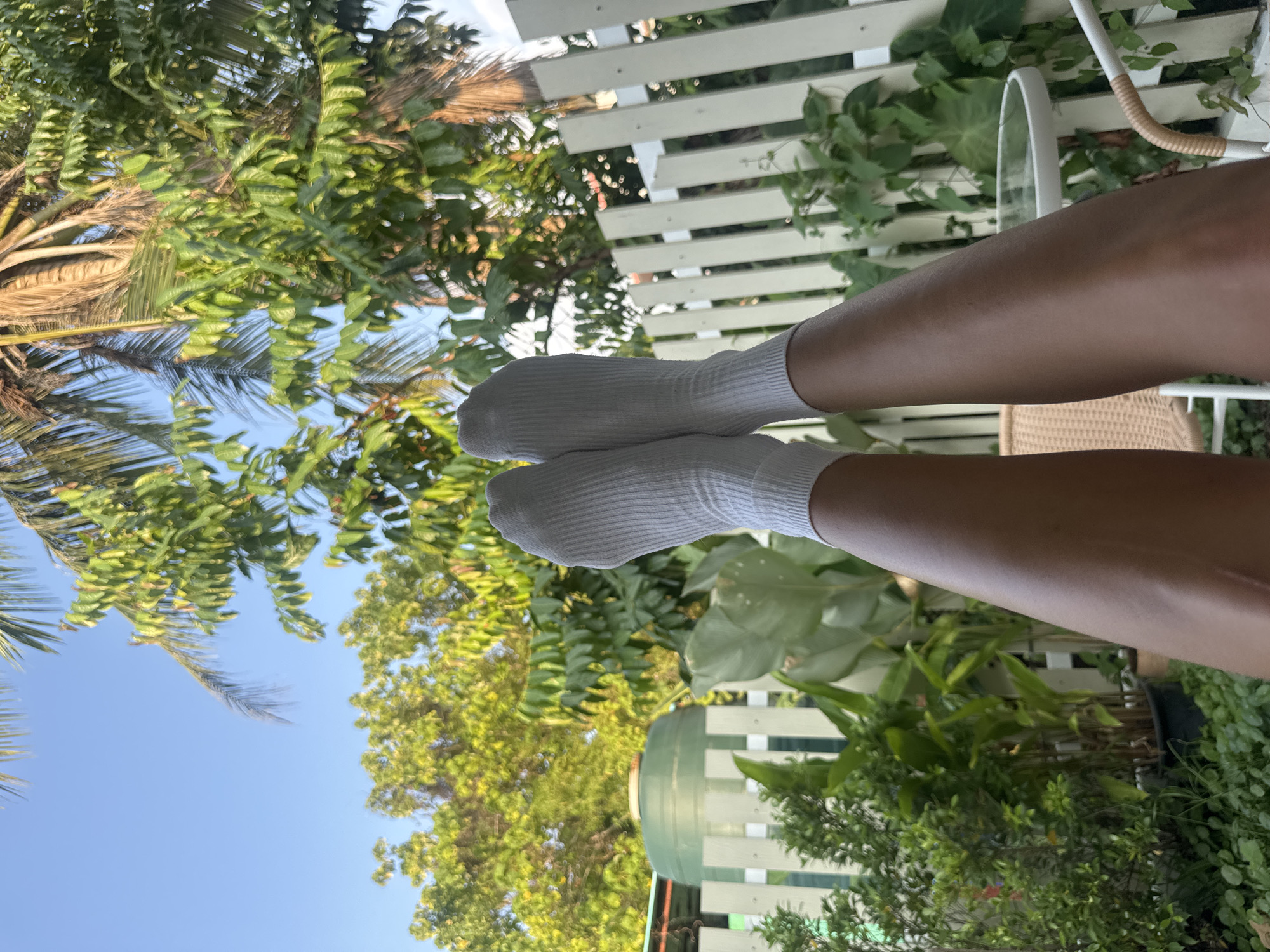The image displays a person's bare legs, clad in white ribbed crew socks, elevated and positioned centrally within an outdoor setting in Choeng Thale, Thailand. The skin on the visible legs is light brown. In the lower frame, portions of a light-colored wicker chair and a white metal table with a circular glass top are present. Behind the elevated legs and foreground furniture, a white picket fence extends horizontally across the midground. Beyond the fence, a dense arrangement of green foliage is visible, including various potted plants and larger trees. To the left of the frame, partially obscured by the fence, a large green cylindrical water tank is situated. The upper portion of the image reveals abundant green tree canopies, including the fronds and trunk of a palm tree, set against a clear, bright blue sky. The scene is illuminated by natural daylight, suggesting an outdoor garden or patio area.