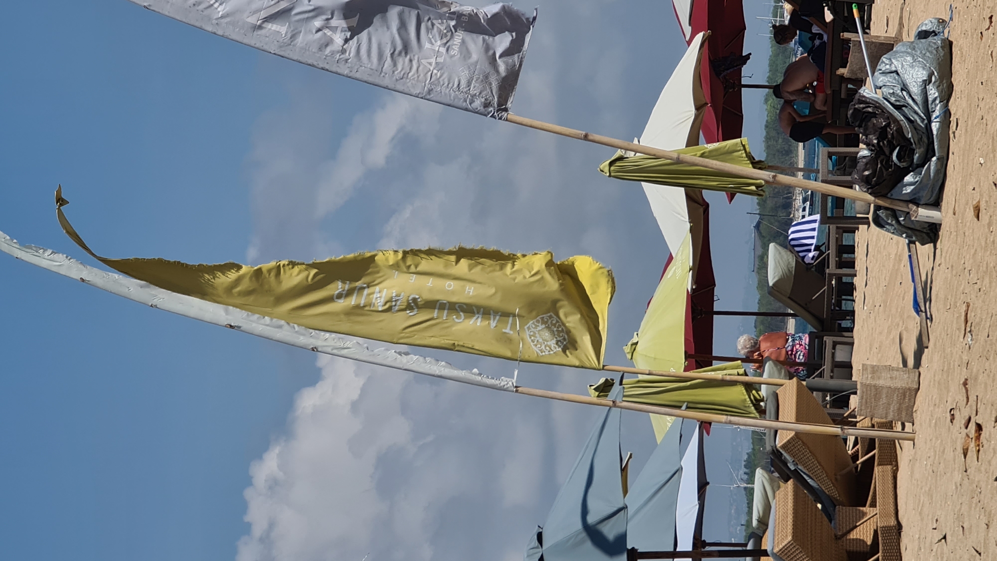 The image depicts a beach scene in Denpasar, Indonesia, likely on a sunny day with some clouds in the sky. In the foreground, two tall flags are planted in the sand. The flag on the left is yellow with white lettering that reads "TAKSU SANUR HOTEL" vertically, along with a circular emblem. The flag on the right is a lighter color, possibly silver or white, with some unreadable text. Behind the flags, several beach umbrellas are open, providing shade. Some of these umbrellas are yellow-green, while others are red and white.
Several beach chairs and loungers are visible, some occupied by people. In the middle ground, a person with white hair is seated on a chair, facing away from the camera, wearing a colorful bathing suit. To the right of this person, two individuals are interacting near loungers, with one appearing to be lying down.
The sand on the beach is a light brown color. In the lower right corner of the image, a large pile of debris wrapped in what appears to be a silver tarp is present. In the background, beyond the umbrellas and loungers, the ocean is visible with some boats anchored in the distance. The overall environment suggests a resort or public beach area.