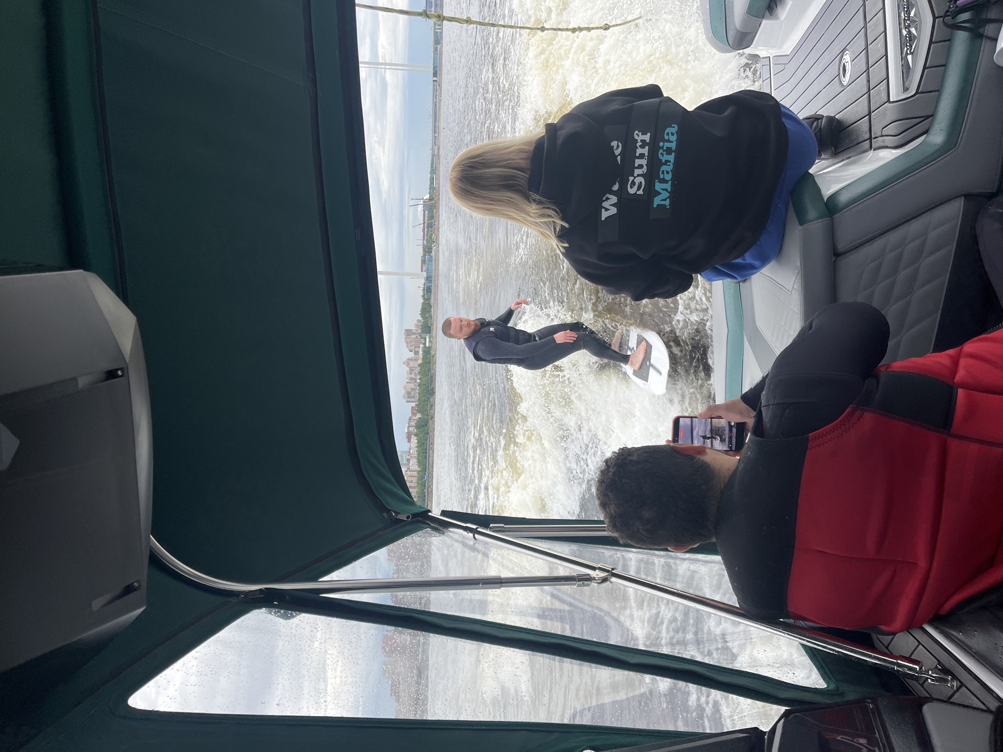 The image shows a scene of wake surfing on a cloudy day in Saint Petersburg, Russia. In the foreground, a person wearing a red and black life vest is seated on a boat, holding a smartphone to record the activity. Their back is to the camera, and their head is turned towards the water. Behind them, another person wearing a black wetsuit is wake surfing on a white board. They are in motion, generating a spray of water.
Further back, a person with blonde hair wearing a black hoodie with the text "Wave Surf Mafia" is standing on the boat. The boat is moving through choppy water, leaving a significant wake. In the background, a cityscape with buildings and industrial structures can be seen, along with a bridge in the distance. The sky is overcast, suggesting a mild or cool temperature. The overall mood appears to be one of recreational activity and enjoyment.