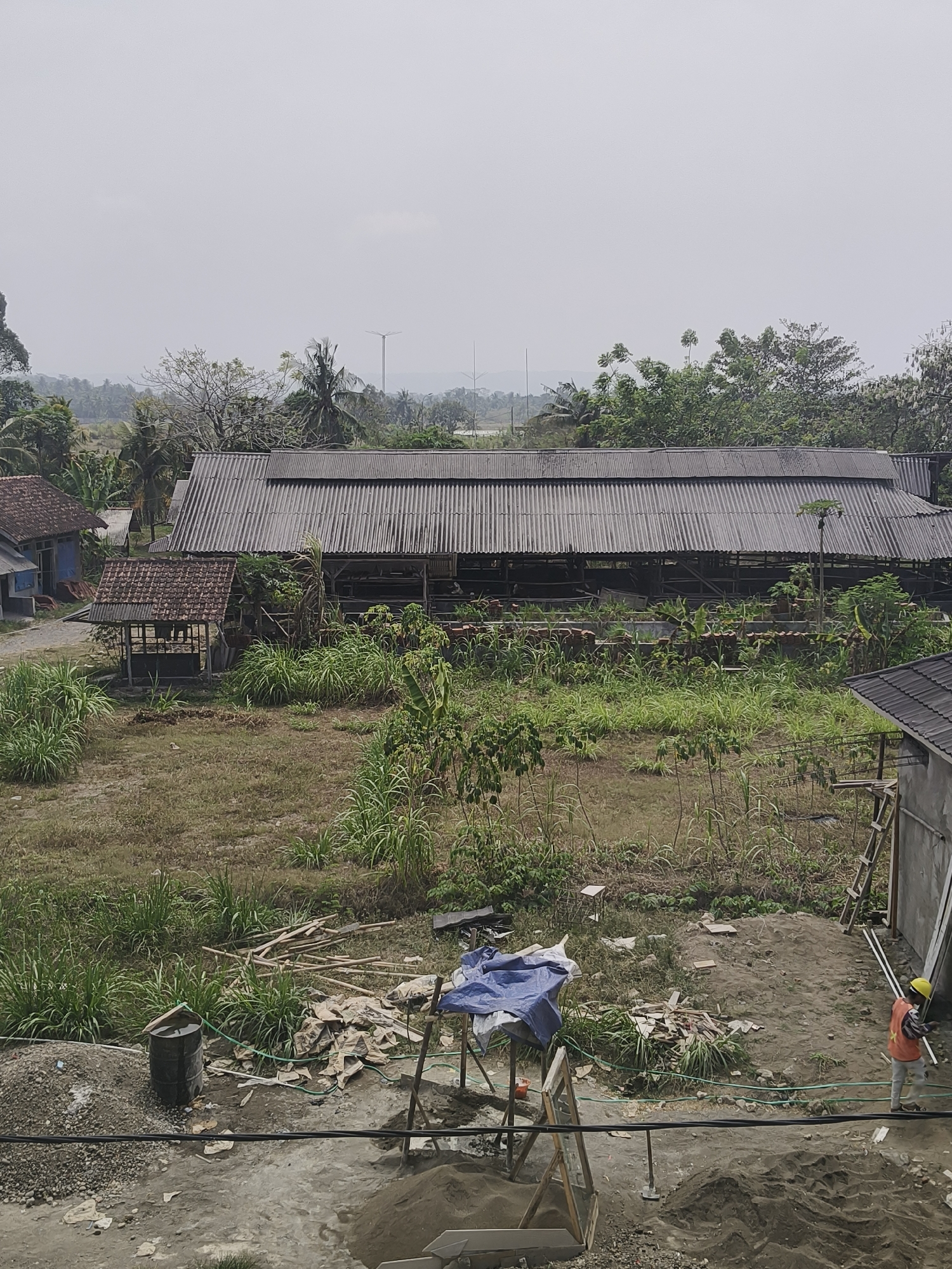 The image shows an outdoor construction site in Desa Cijulang, Indonesia. In the foreground, a pile of gravel and sand is visible, along with a black metal drum and some scattered construction debris. A construction worker wearing a yellow hard hat and an orange vest is walking across the scene. In the middle ground, there is a large, corrugated metal roof structure, possibly a barn or warehouse, with an open-sided section underneath. A smaller, tiled-roof structure is situated to the left of the large building. The surrounding area is overgrown with tall grass and trees. In the background, more trees and vegetation are visible, with a hazy sky overhead. The overall scene depicts an active construction area within a rural setting.