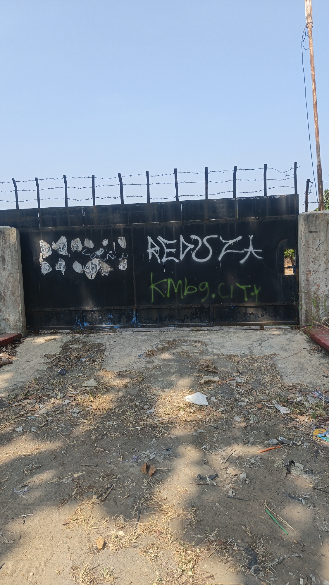 The image shows a black metal gate with barbed wire on top, set against a clear blue sky. The gate is covered in graffiti. On the left side of the gate, there are patches of white and grey spray paint and stickers. In the center and to the right, white graffiti reads "REDUSZA" and green graffiti below it reads "Kmbg.CITY". The gate is supported by concrete pillars on either side. In front of the gate, the ground is rough and unpaved, covered with dirt, dry grass, and scattered debris, including plastic bags and wrappers. A rusty metal pole stands to the right of the gate, with a wire attached to its top. The scene appears to be an entrance to an industrial or abandoned area. The location is Jekulo, Indonesia.