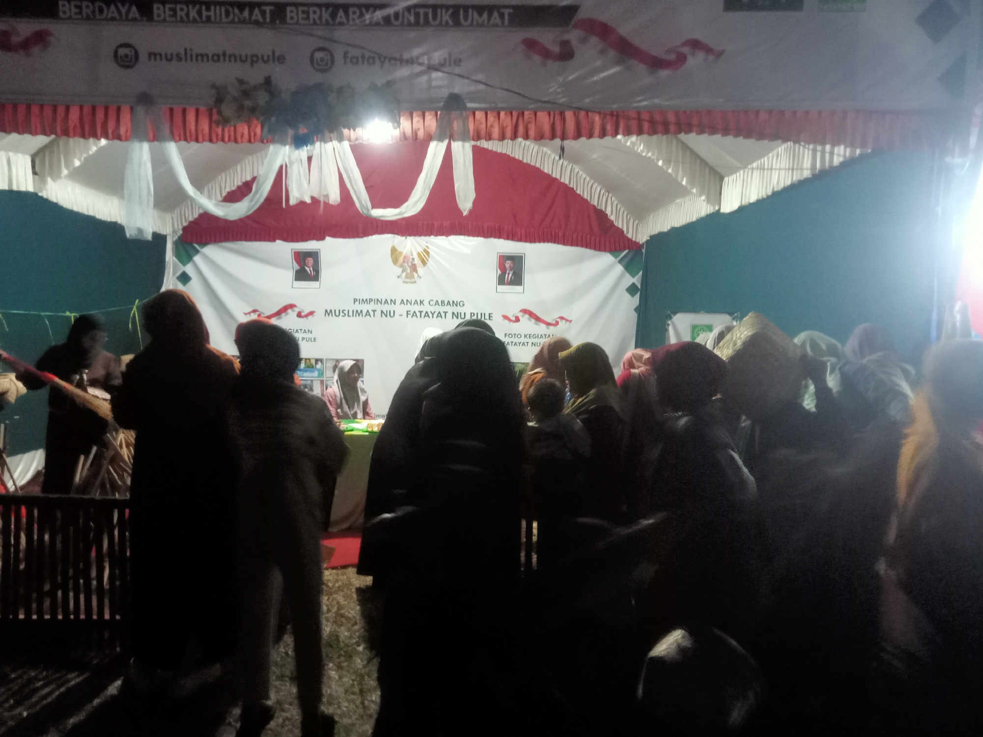 The image depicts an outdoor event at night in Pule, Indonesia, likely a cultural or religious gathering. A tent structure with red and white draped fabric serves as a backdrop. A banner displays the text "BERDAYA BERKHIDMAT. BERKARYA UNTUK UMAT" and social media handles "@muslimatnpule" and "@fatayatpule". Another banner in the background features images of what appear to be political figures and the Garuda Pancasila symbol, along with text that seems to indicate "PIMPINAN ANAK CABANG MUSLIMAT NU - FATAYAT NU PULE". A group of people, mostly silhouetted and wearing headscarves, are gathered in front of the stage area, some holding what looks like a broom or stick. The lighting is dim, with artificial lights illuminating the scene. The ground appears to be covered in grass or straw. The overall atmosphere suggests a community event, possibly a religious or political rally associated with Muslimat NU and Fatayat NU, organizations connected to Nahdlatul Ulama in Indonesia.