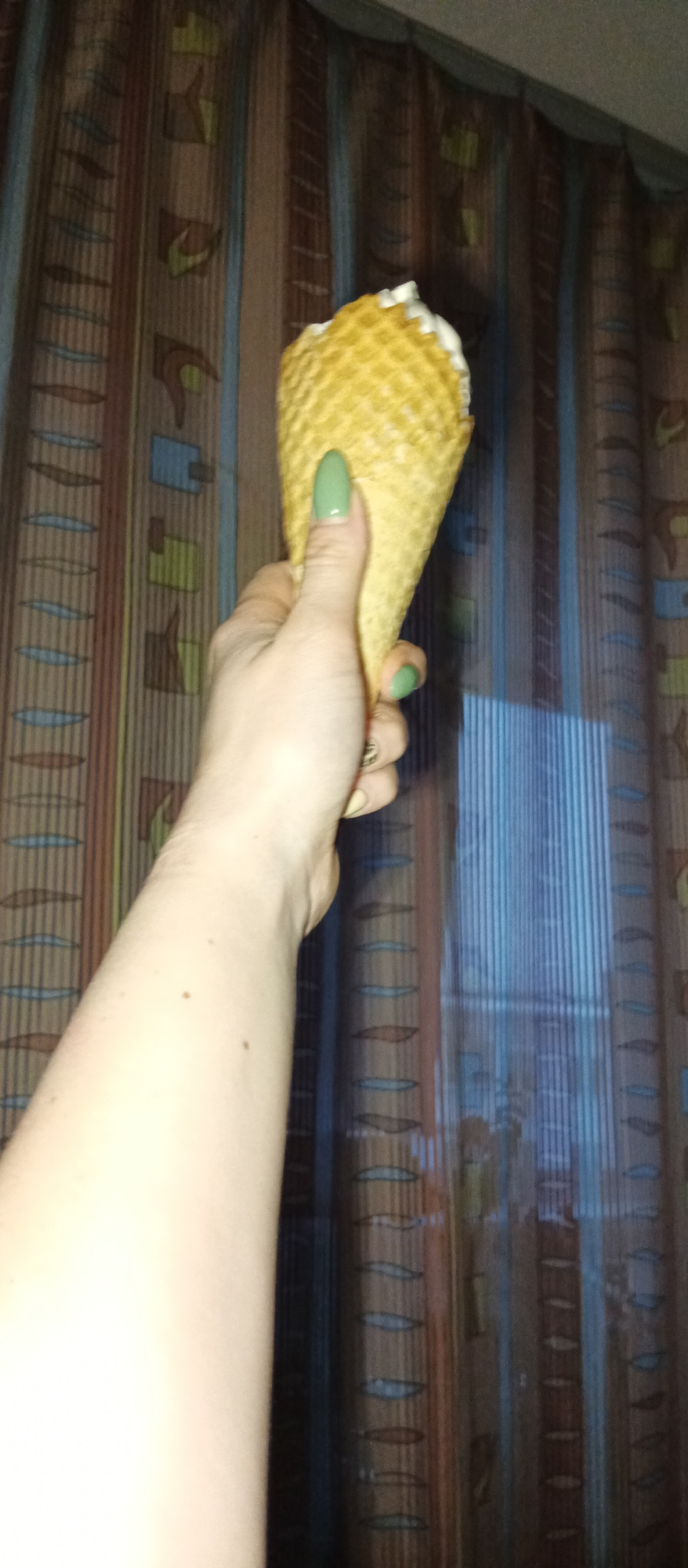 A person's arm and hand are holding an ice cream cone filled with a white substance, possibly ice cream, against a backdrop of curtains with a vertical striped pattern. The person has pale skin and is wearing green nail polish. The ice cream cone is golden-brown with a waffle pattern. The location is unknown.
