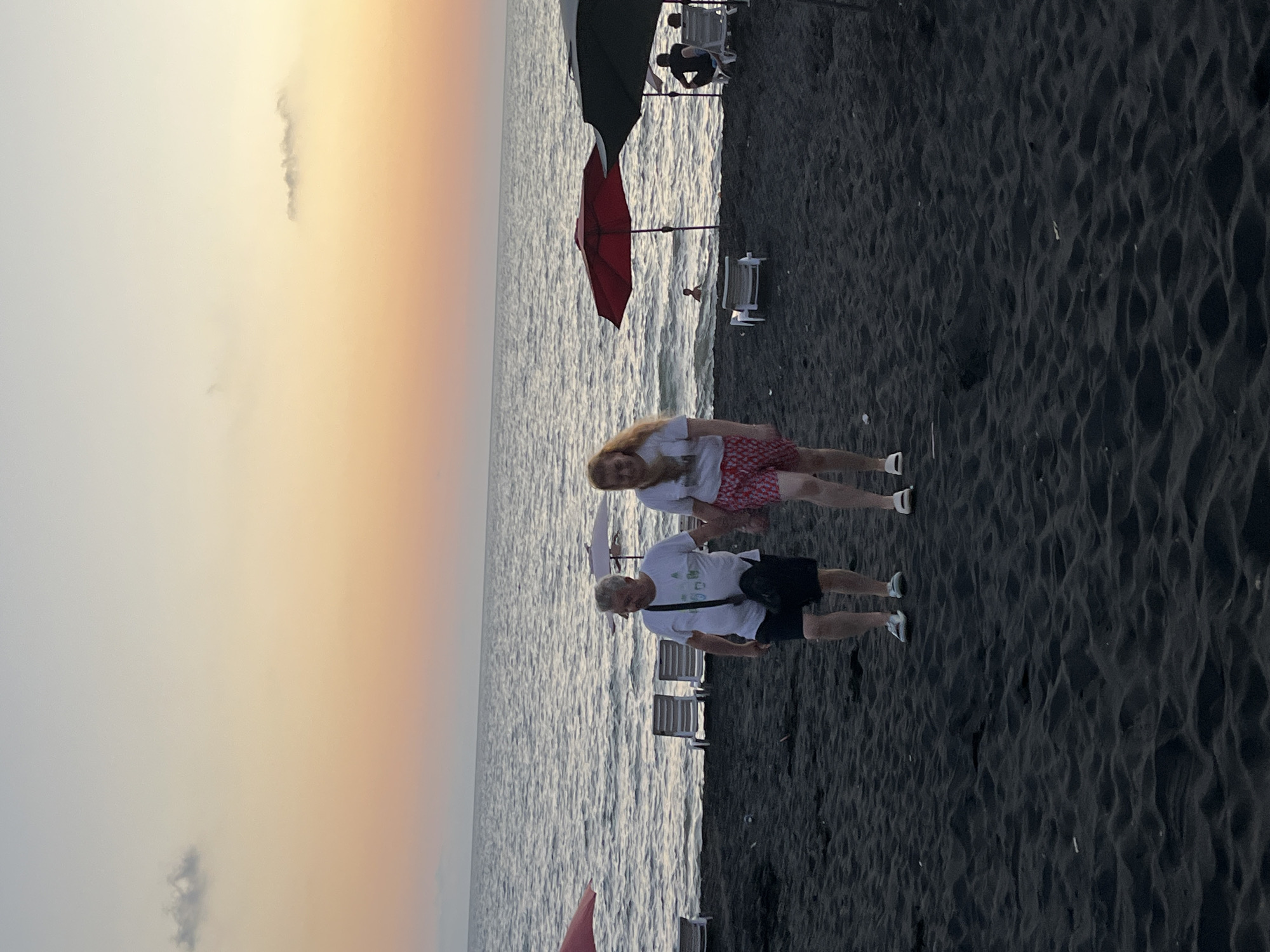 The image captures a sunset scene on the black sand beach of Ureki, Georgia. Two individuals, an older man and a younger woman, are walking hand-in-hand across the dark sand towards the viewer. The man wears a white t-shirt and dark shorts, with a black bag slung across his shoulder. The woman is dressed in a white t-shirt and red patterned shorts.
The sea stretches out behind them, its surface rippled by gentle waves under a sky painted with soft hues of orange and yellow, indicating dusk. Along the shoreline, several beach chairs and umbrellas are visible, some occupied by other beachgoers. A red umbrella stands out near the water's edge, with a person swimming in the sea nearby. Further down the beach, dark umbrellas and chairs suggest more people are enjoying the evening. The overall atmosphere is one of a peaceful end to a day at the beach.