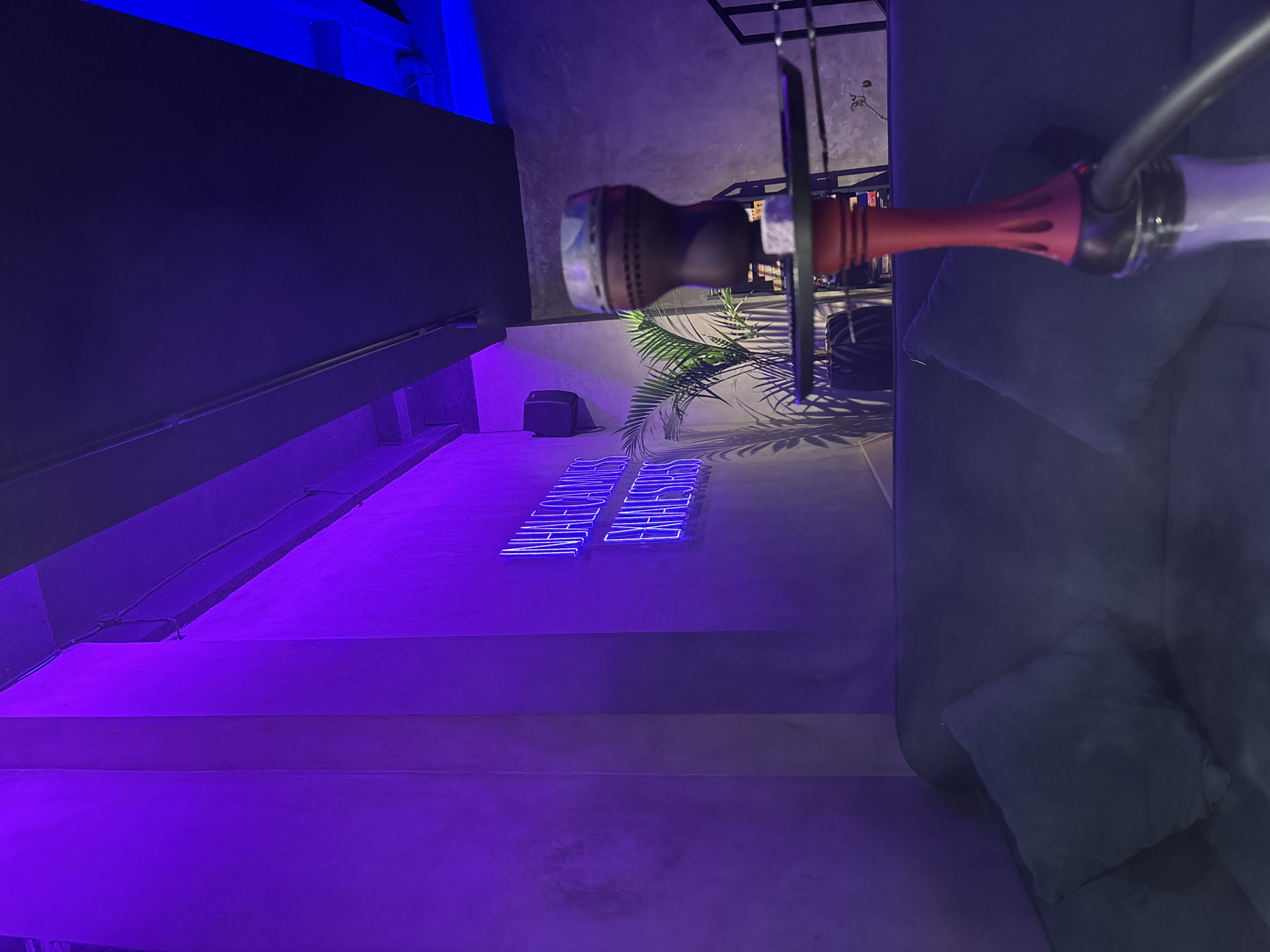 The image shows a dimly lit interior, possibly a bar or lounge in Kuta Selatan, Indonesia. The lighting is primarily blue and purple, creating a moody atmosphere. A hookah is prominently featured, its bowl and hose extending into the frame. The background includes plants, a dark speaker, and neon lights spelling out words. The floor appears to be concrete or a similar material, with steps leading up to a platform. Part of a sofa is visible, suggesting a seating area. There is no evidence of people.