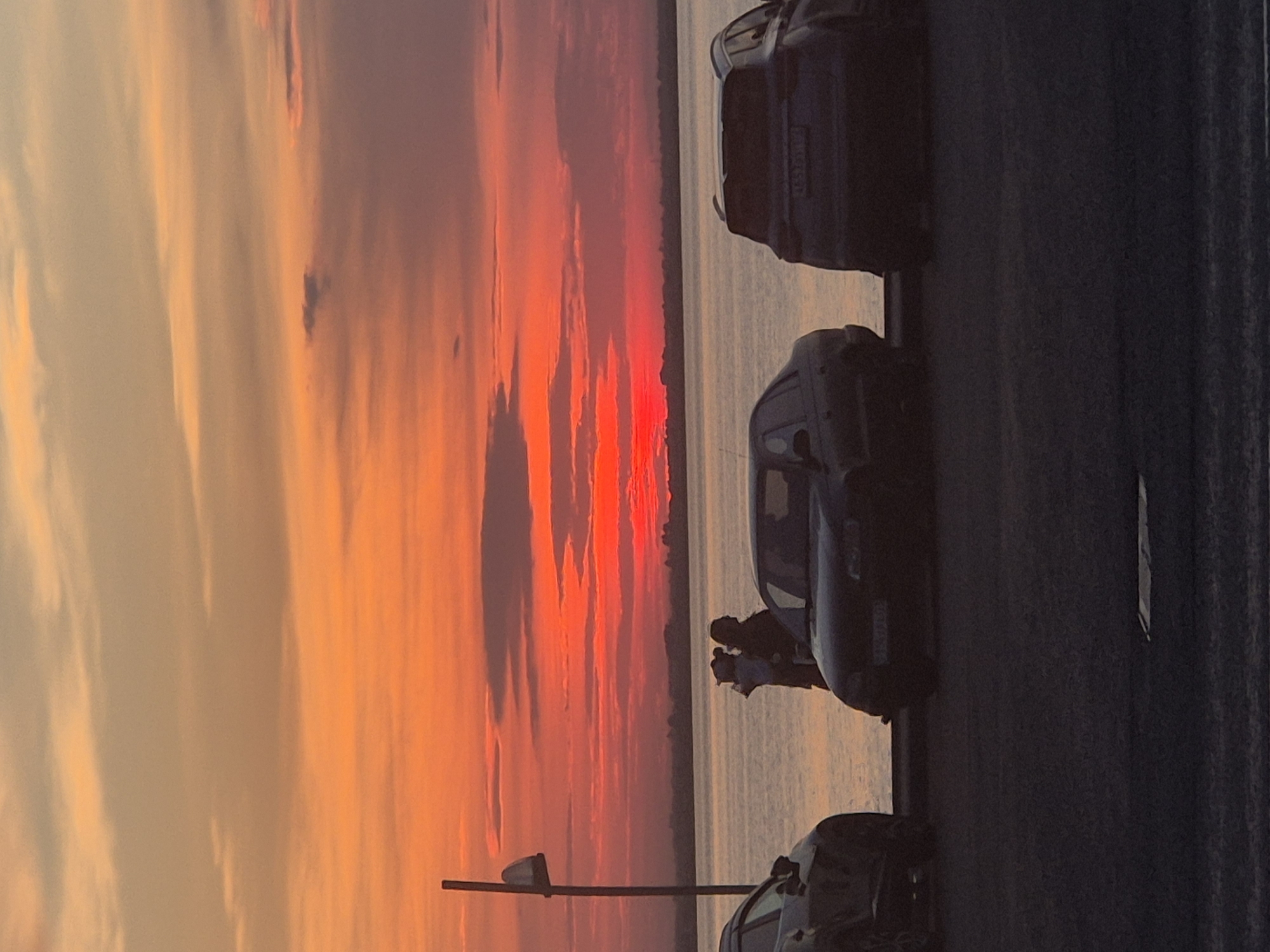 An outdoor, low-angle, vertical shot captures a sunset over a body of water. In the foreground, a paved surface with faint white lines suggests a road or parking area. Three cars are parked, one on the left facing right, one in the center facing right, and one on the right facing left. The car on the left is partially obscured. The center car is black, and the car on the right is a dark SUV.
Between the center and right cars, silhouetted against the water and sky, are two people standing. One person appears to be holding a child. In the middle ground, a wide expanse of water reflects the sky's colors. A thin, dark horizontal line of land is visible on the horizon, separating the water from the sky.
The sky is dominated by a vibrant sunset, transitioning from deep oranges and reds near the horizon to lighter oranges and yellows higher up, with streaks of pink and pale yellow clouds. A single, tall, thin vertical pole with a lamp at the top stands on the left side of the image, between the left car and the water. The scene is devoid of any specific landmarks that would pinpoint it to Kokshetau, Kazakhstan; it is a generic scene of a sunset by a body of water with parked cars and people.